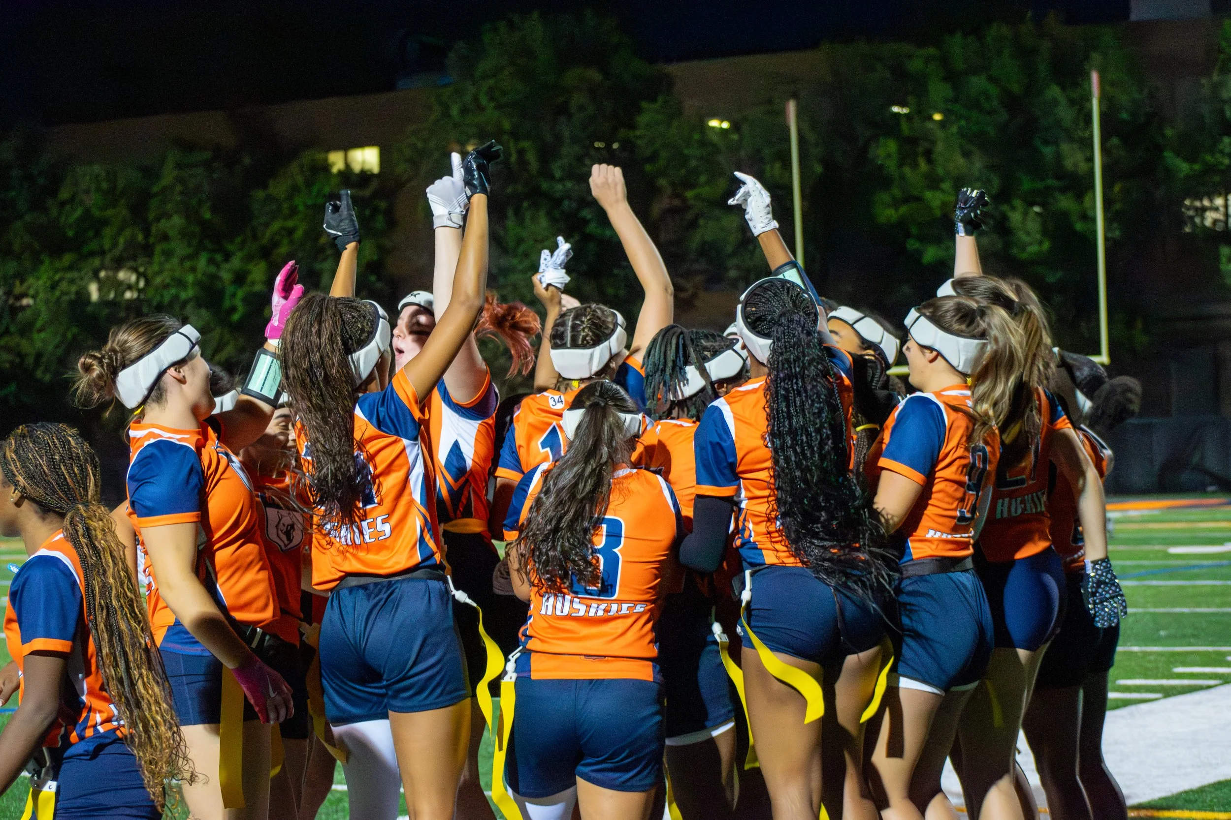 Group of female football players in orange and blue uniforms celebrating on the field at night.