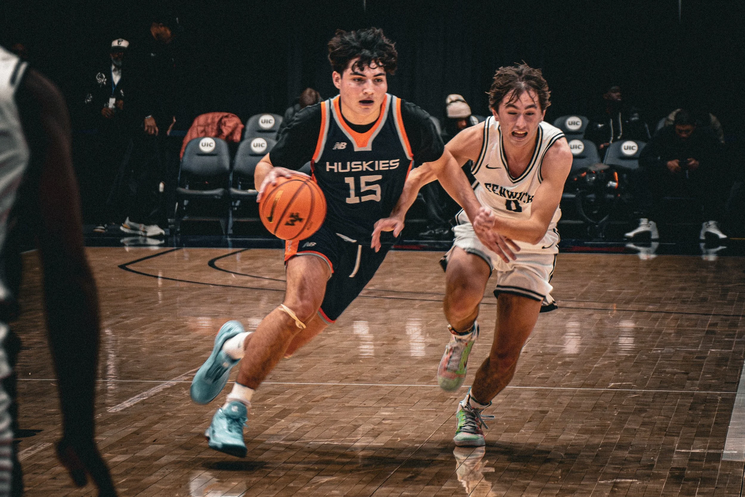 Two young male basketball players competing for the ball on a court, one in a black and orange uniform and the other in a white uniform. The player in black is dribbling, while the player in white is trying to reach for the ball.