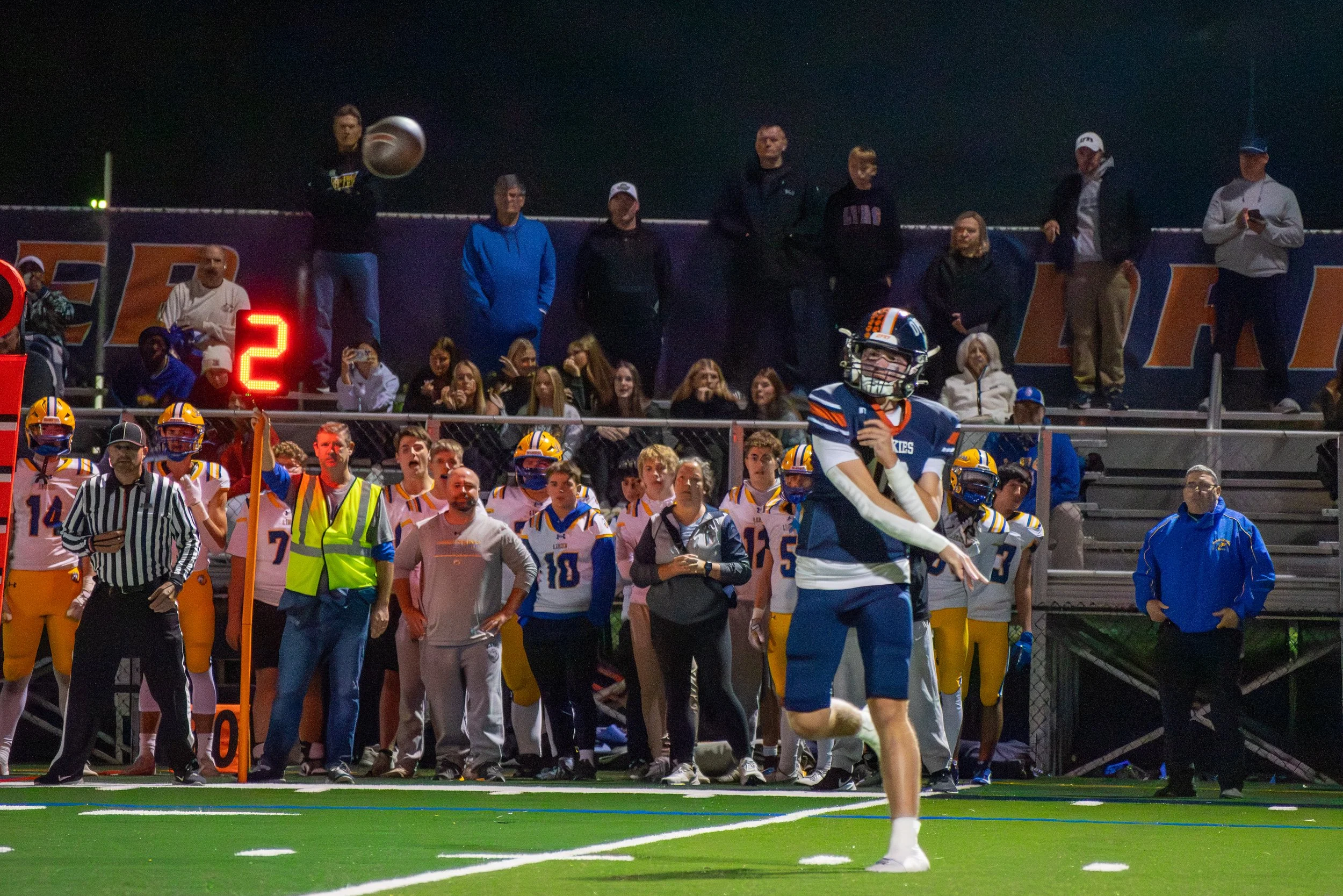 A football player in a blue and white uniform on the field with teammates and coaches on the sideline, with spectators watching from the bleachers during a nighttime game.