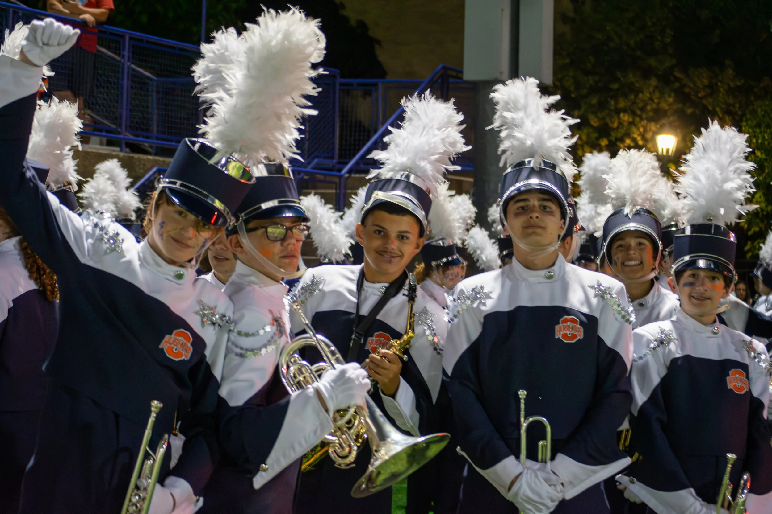 A group of young marching band members wearing black and white uniforms with blue accents, black hats with white plumes, and face paint, standing outdoors at night. Some are holding musical instruments.
