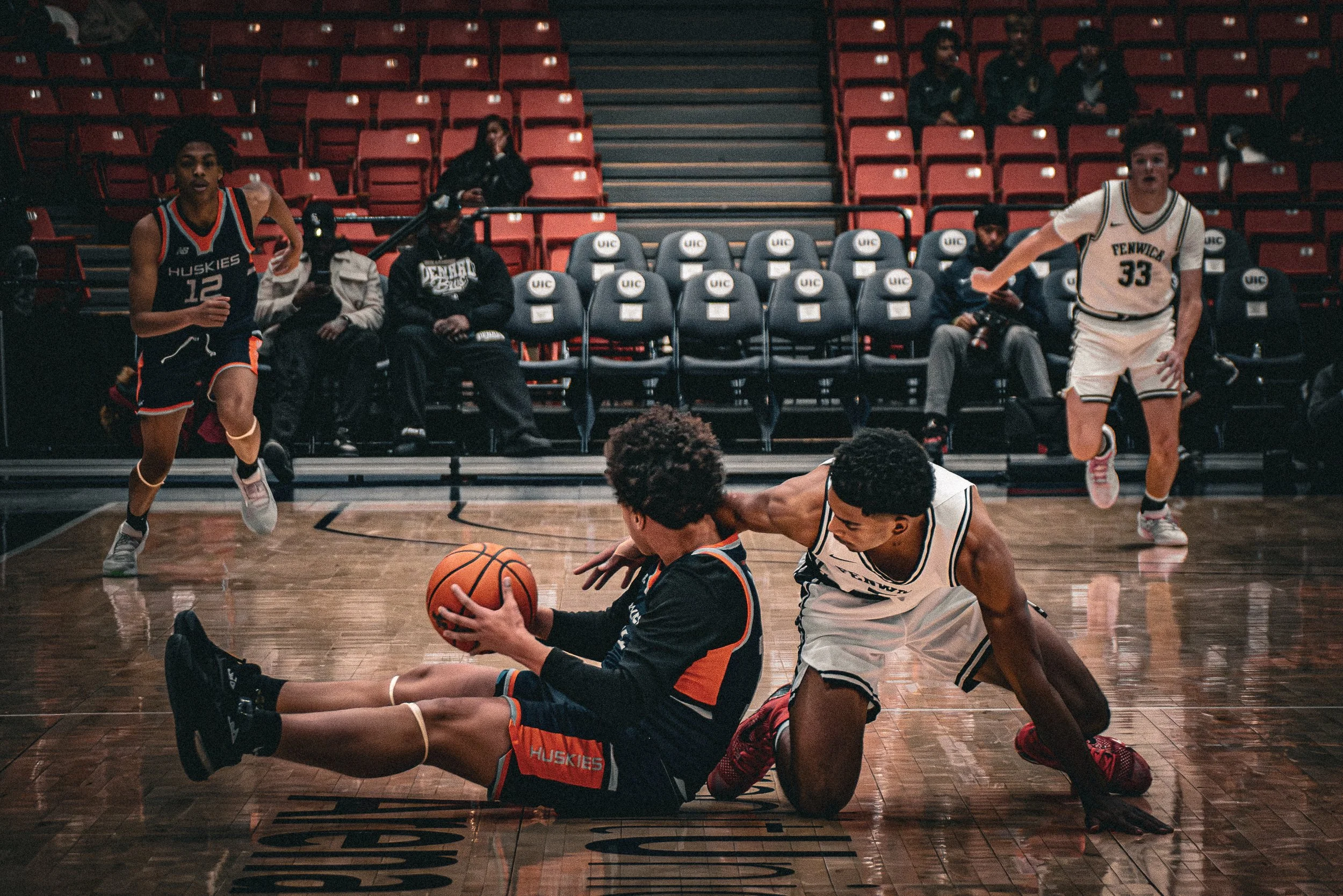 Two young male basketball players competing for the ball on the court, with others running in the background and spectators seated in the stands.