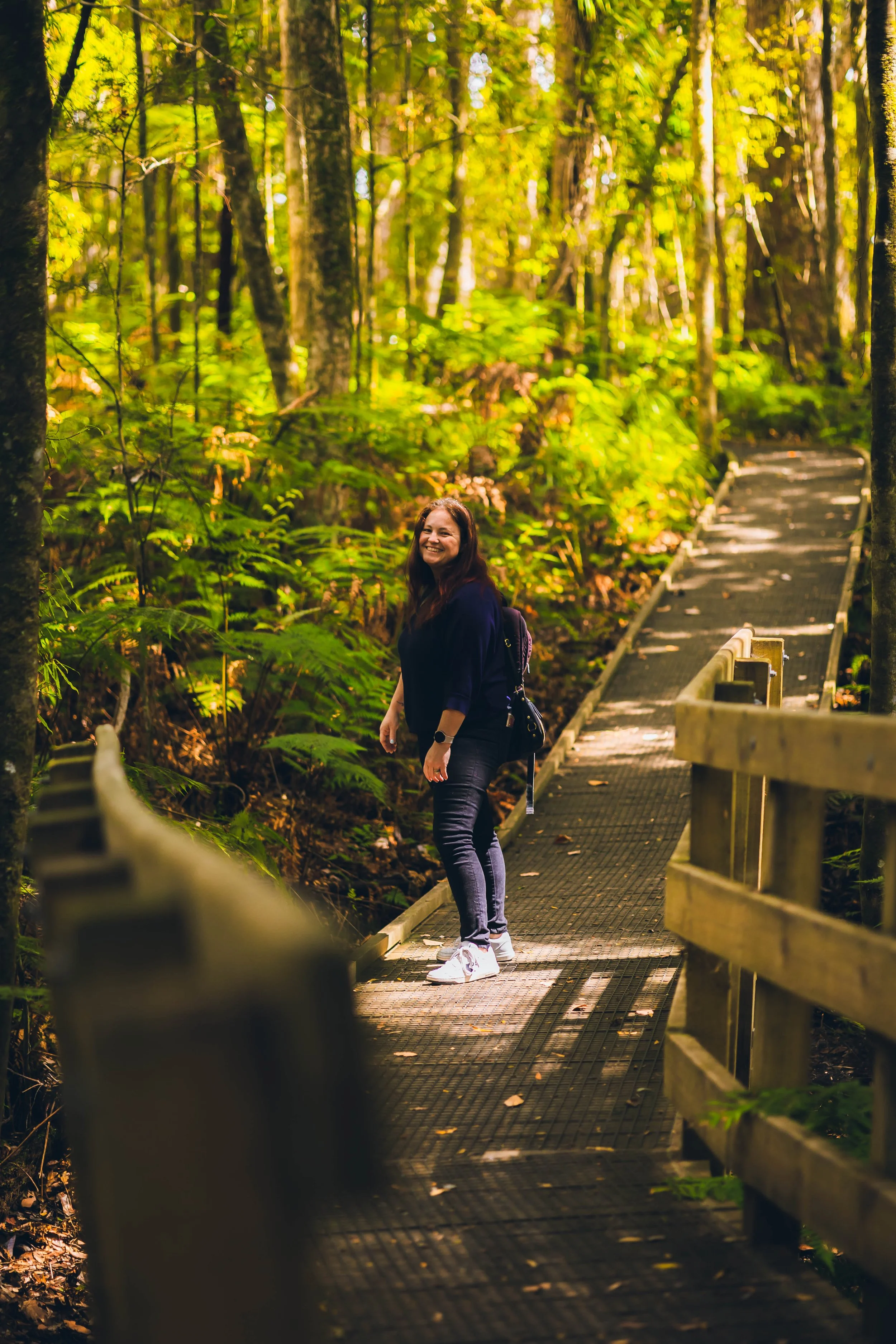 A woman with long dark hair, dressed in dark clothing, stands on a narrow, elevated forest trail with a handrail on one side, smiling amidst lush green trees and foliage in a forest setting during daytime.