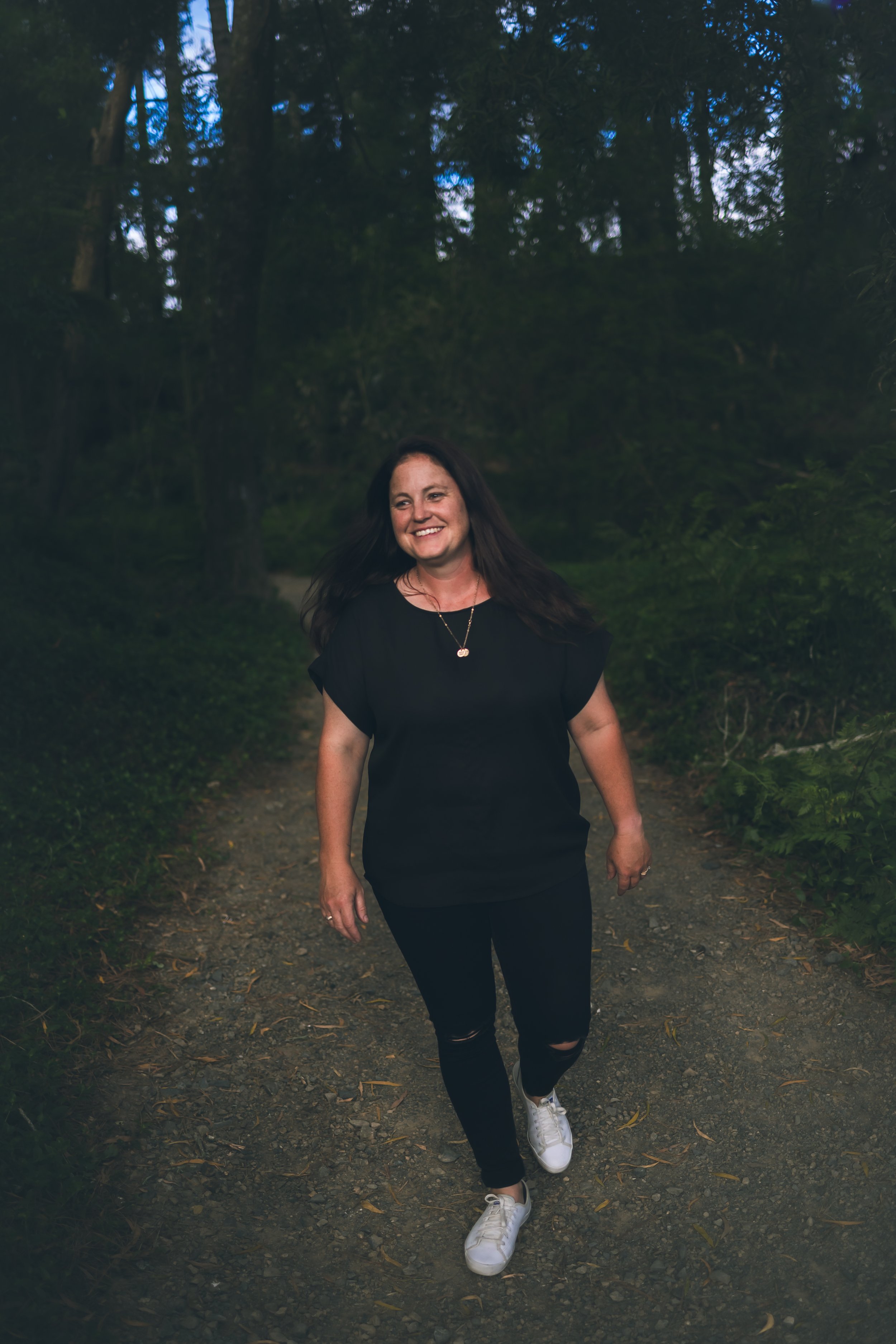 A woman walking on a forest trail during dusk, smiling and wearing a black shirt, ripped black jeans, and white sneakers.