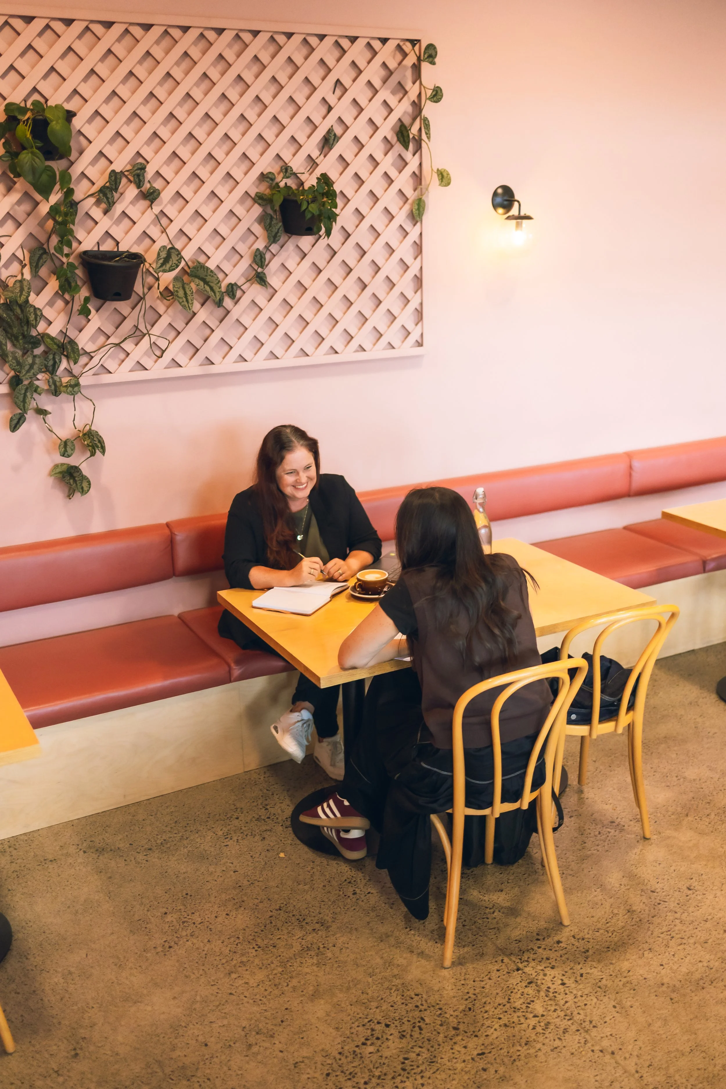Two women sitting at a wooden table in a coffee shop, talking and smiling. One woman has long dark hair, and the other has shoulder-length dark hair. The decor includes a pink wall with a lattice panel with hanging green plants and a wall lamp.