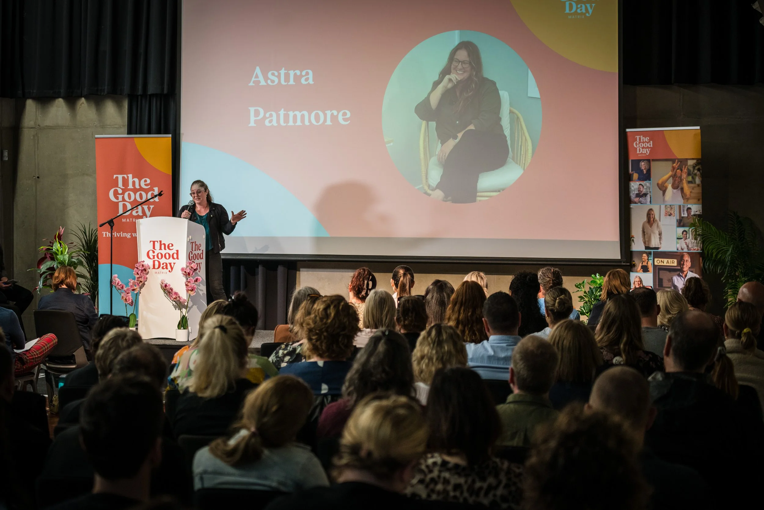 A woman presenting on stage at a conference titled 'The Good Day.' She stands behind a podium with pink flowers and a sign reading 'The Good Day.' The large screen behind her shows her photo and the name 'Astra Patmore'.