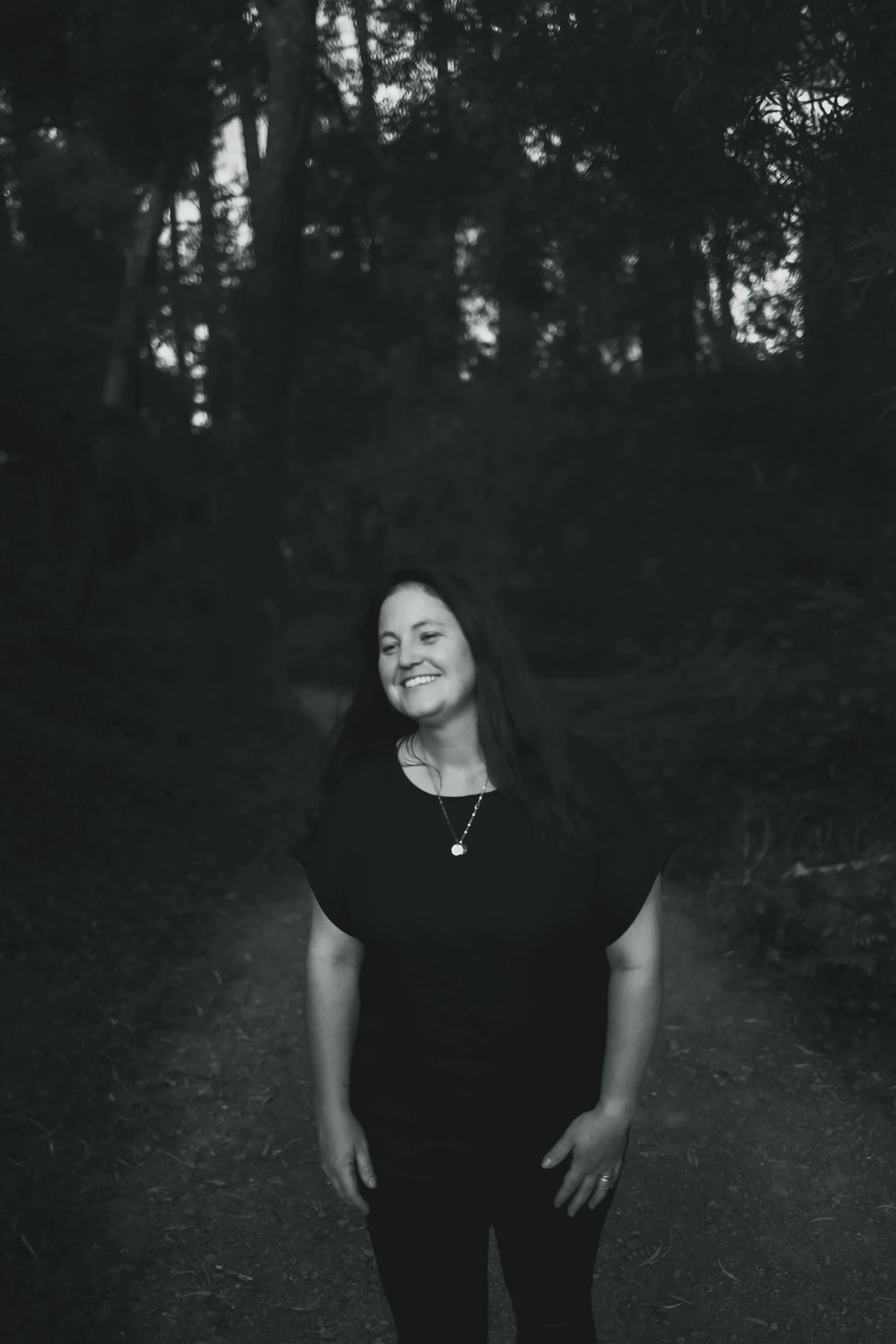 A woman smiling and standing outdoors in a wooded area, wearing a black top and pants, with her hands resting on her thighs.