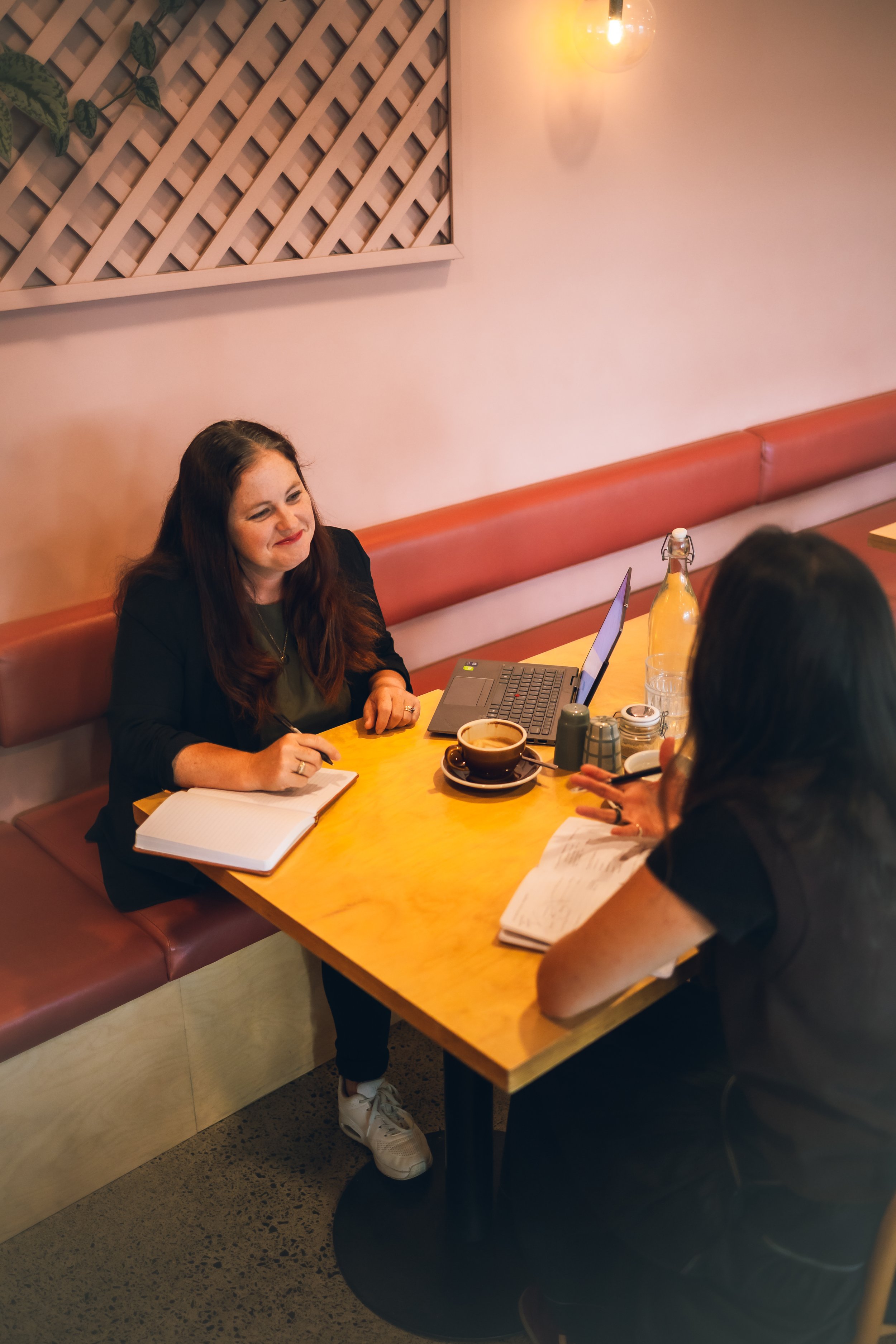 Two women sitting across each other at a wooden table in a cozy cafe, engaged in a discussion with notebooks, laptop, and coffee cups on the table.