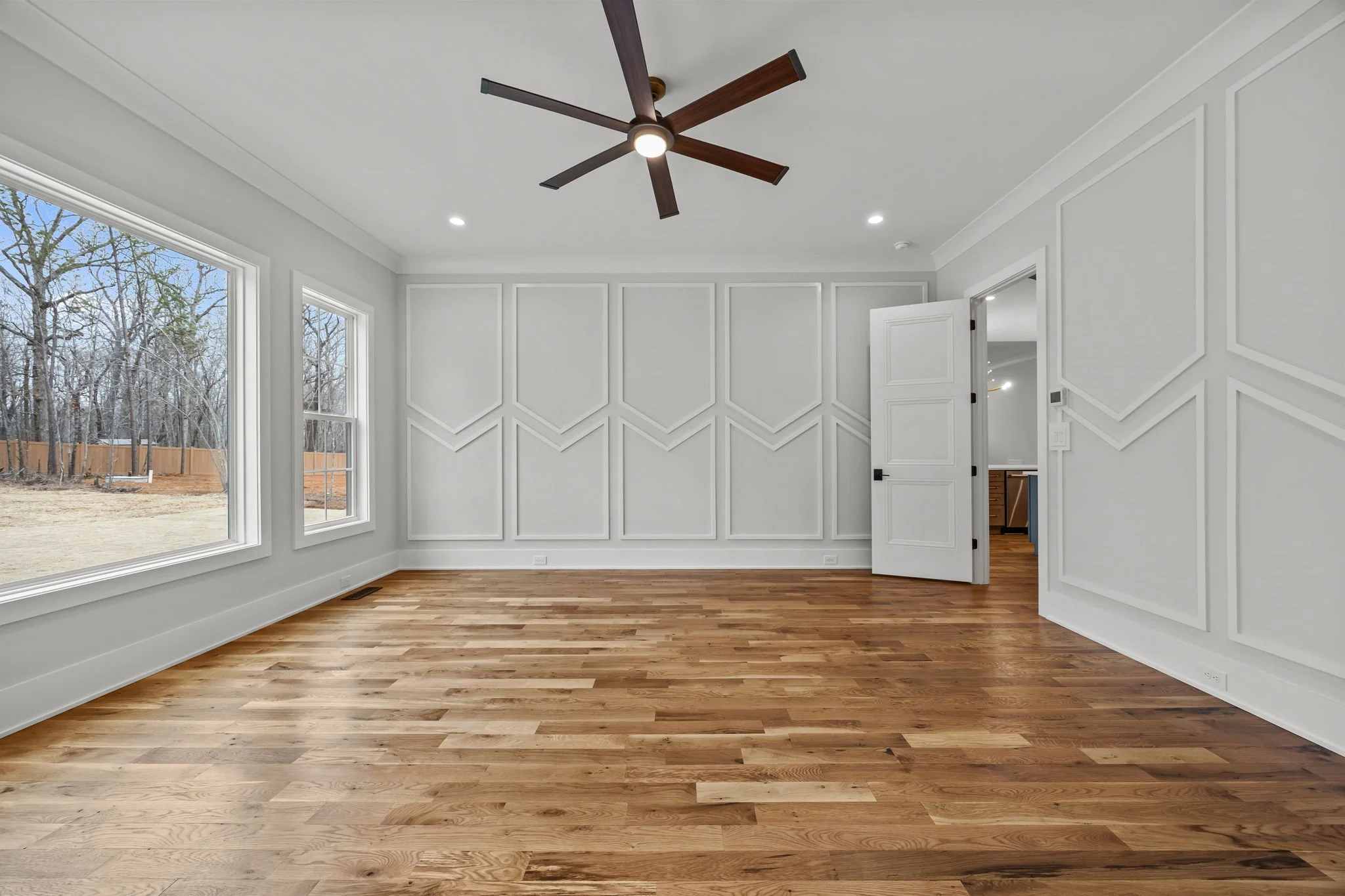 Spacious primary bedroom with large windows, hardwood floors, modern ceiling fan, and decorative wall molding.