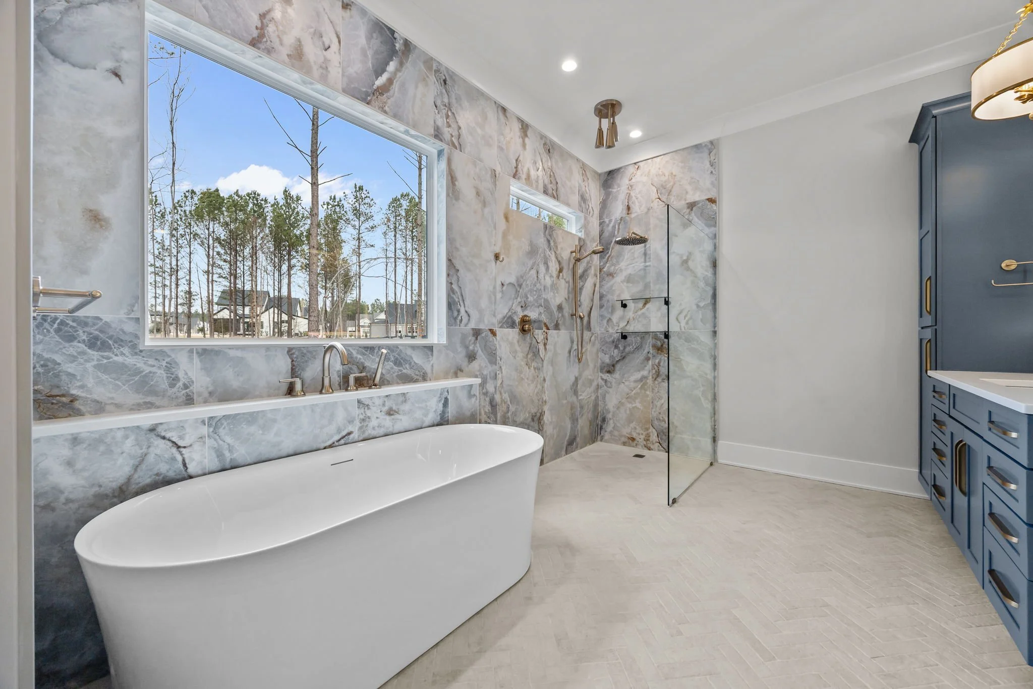 Bright primary bathroom with freestanding soaking tub, floor-to-ceiling marble tile accent wall, glass walk-in shower, large window, and brass fixtures.