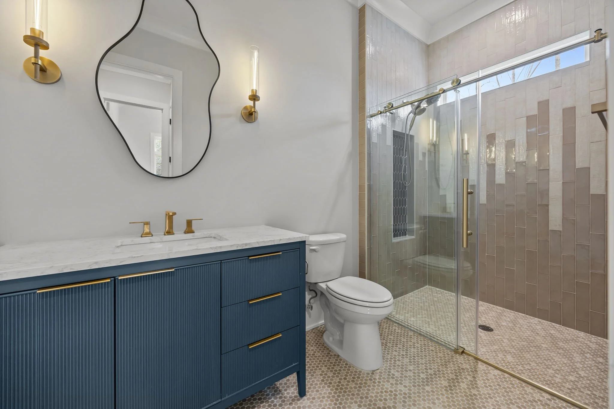 Bathroom vanity with blue cabinetry, brass hardware, quartz countertop, modern mirror, and glass walk-in shower with tile surround.