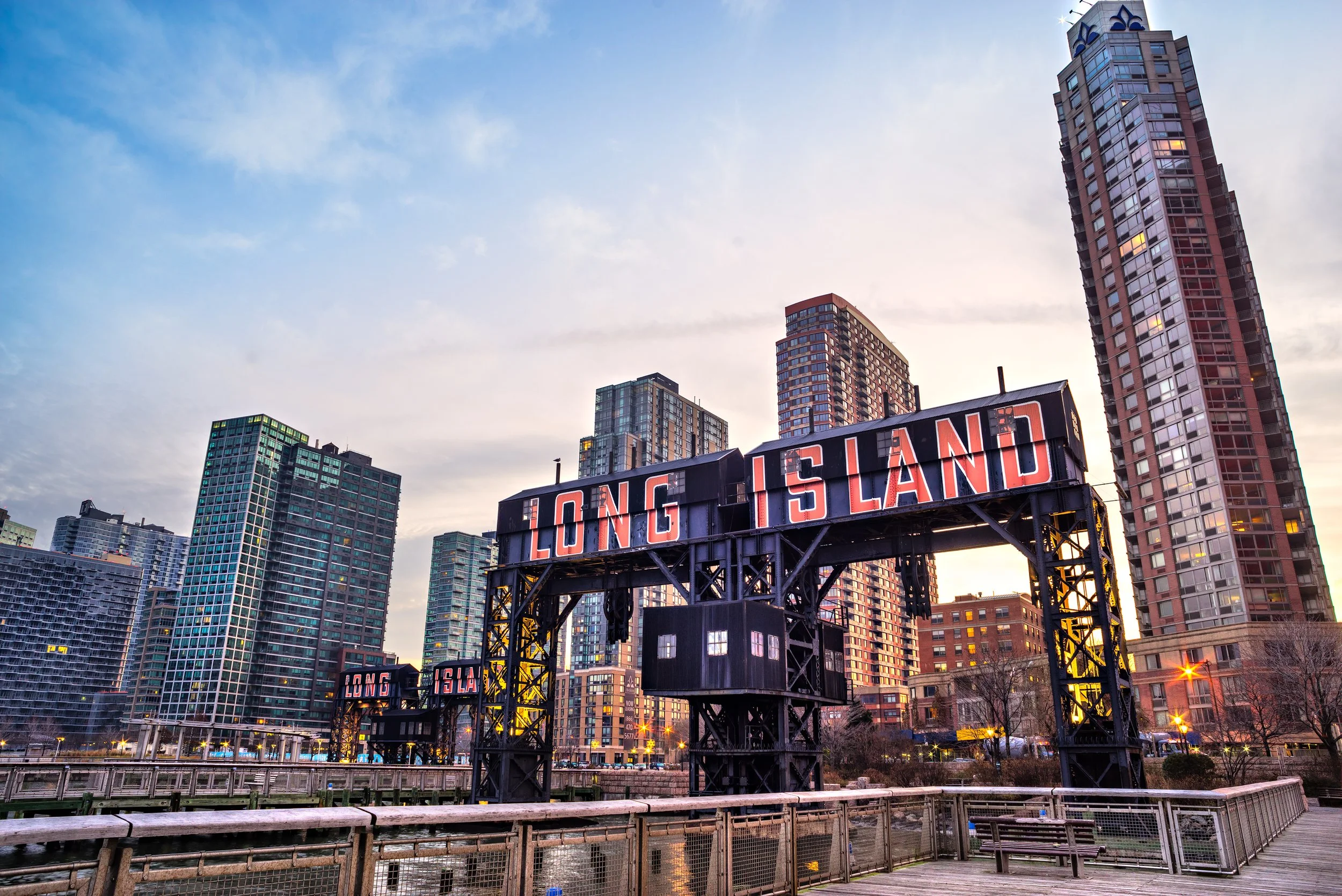 View of a city skyline with tall buildings at sunset and a billboard that reads 'LONG ISLAND' in the foreground.