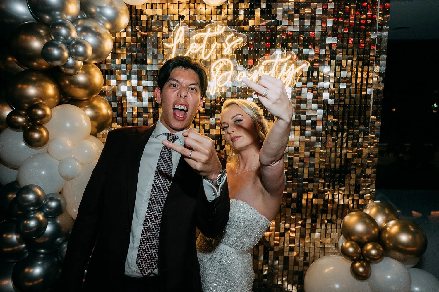 Fun, edgy portrait of the couple flashing their wedding rings and making rock-on gestures in front of a shimmer wall.
