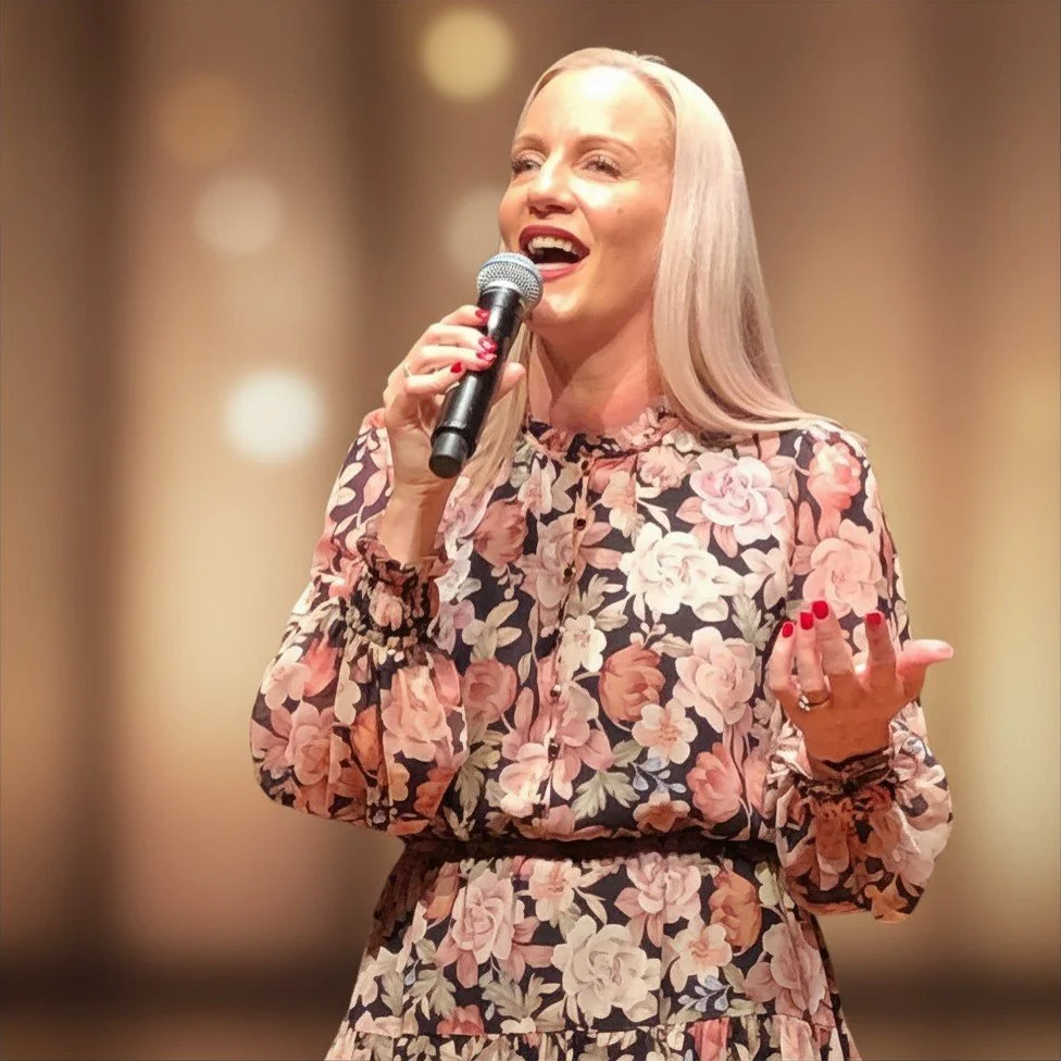 A woman with long blonde hair singing into a microphone, wearing a floral dress with a black background and pink and cream flowers, with a blurred warm-toned background.