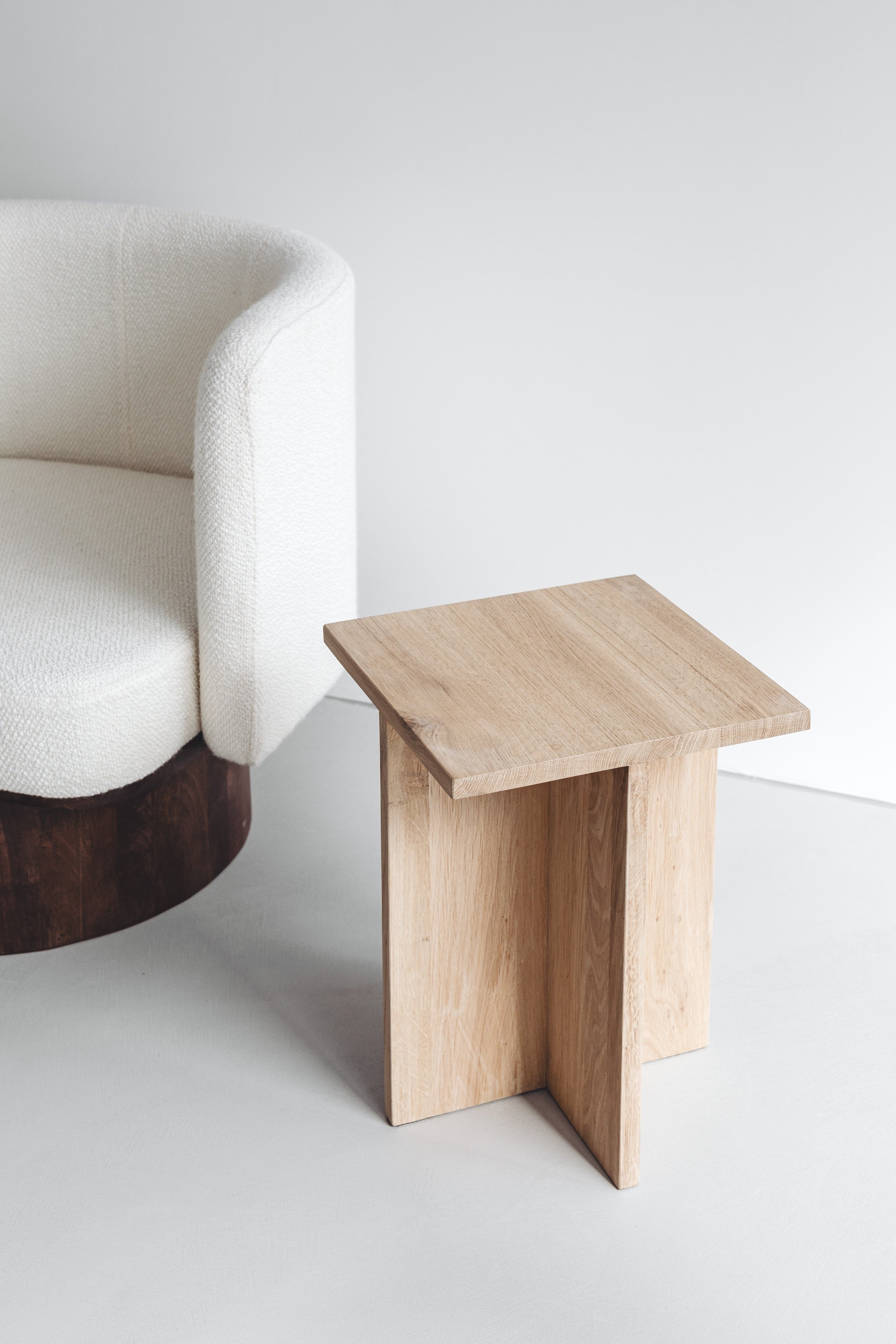 A white upholstered armchair with a dark wooden base next to a small wooden side table on a white floor against a white wall.