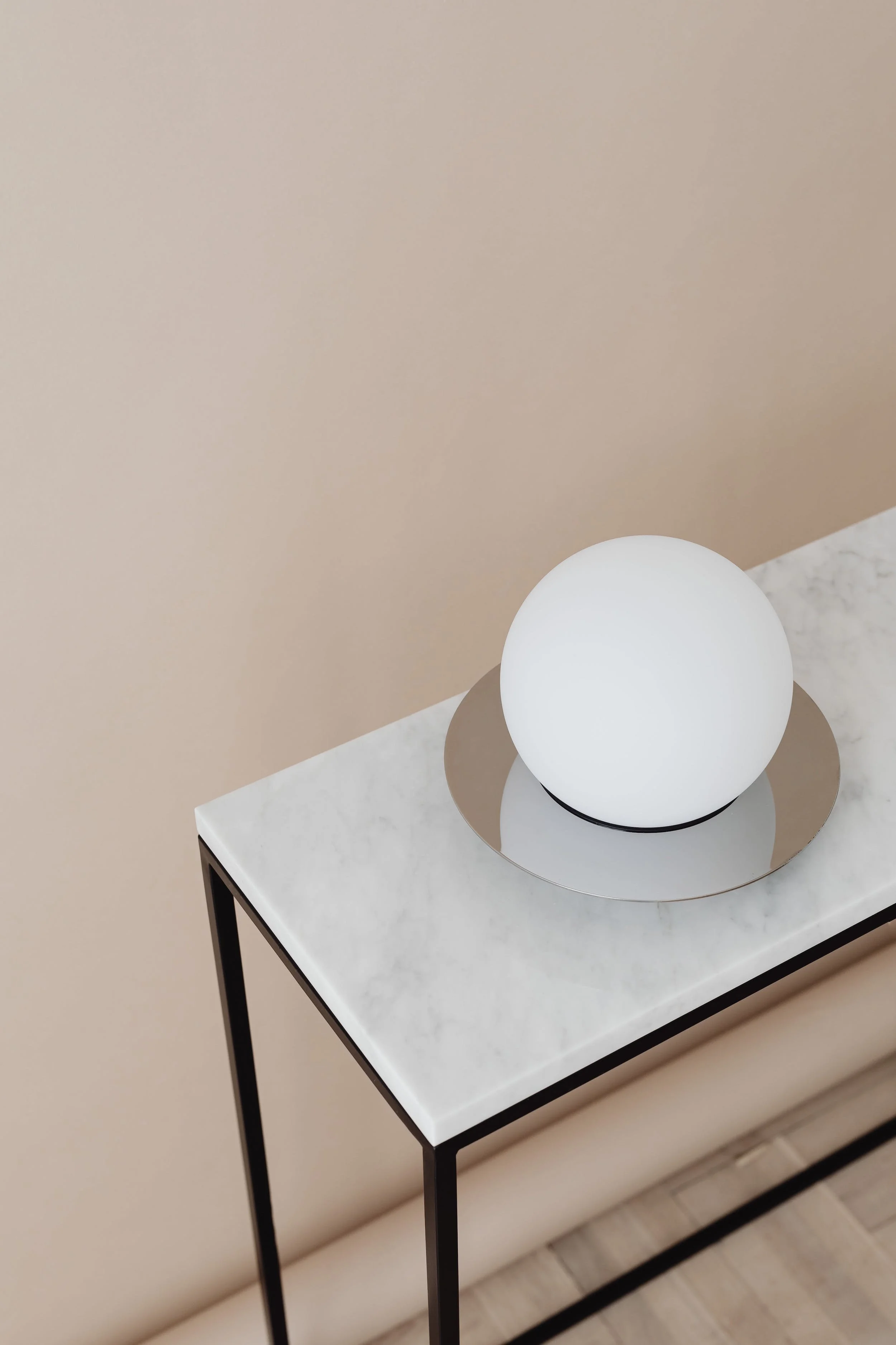 A modern table with a white marble top and black metal legs, holding a spherical white lamp on a mirrored base, against a light beige wall.