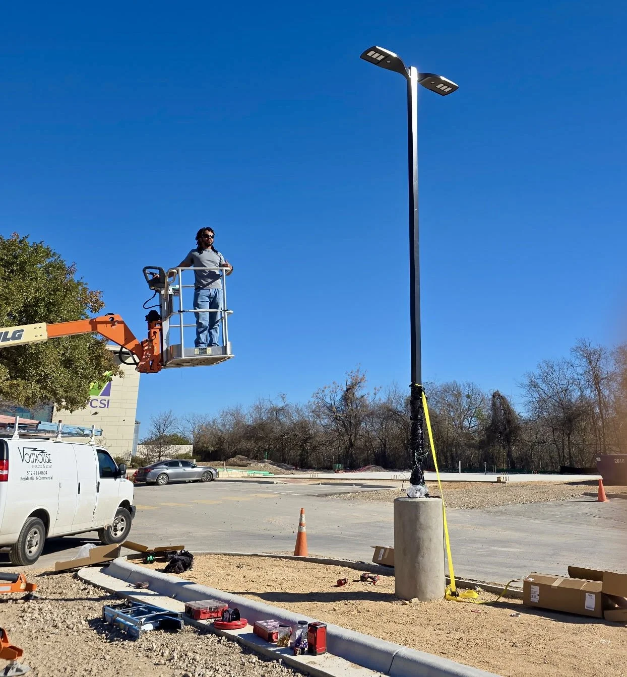 A worker in a bucket lift installing or maintaining a tall streetlight in a parking lot on a sunny day. There are tools and boxes on the ground, orange traffic cones, and parked cars in the background.