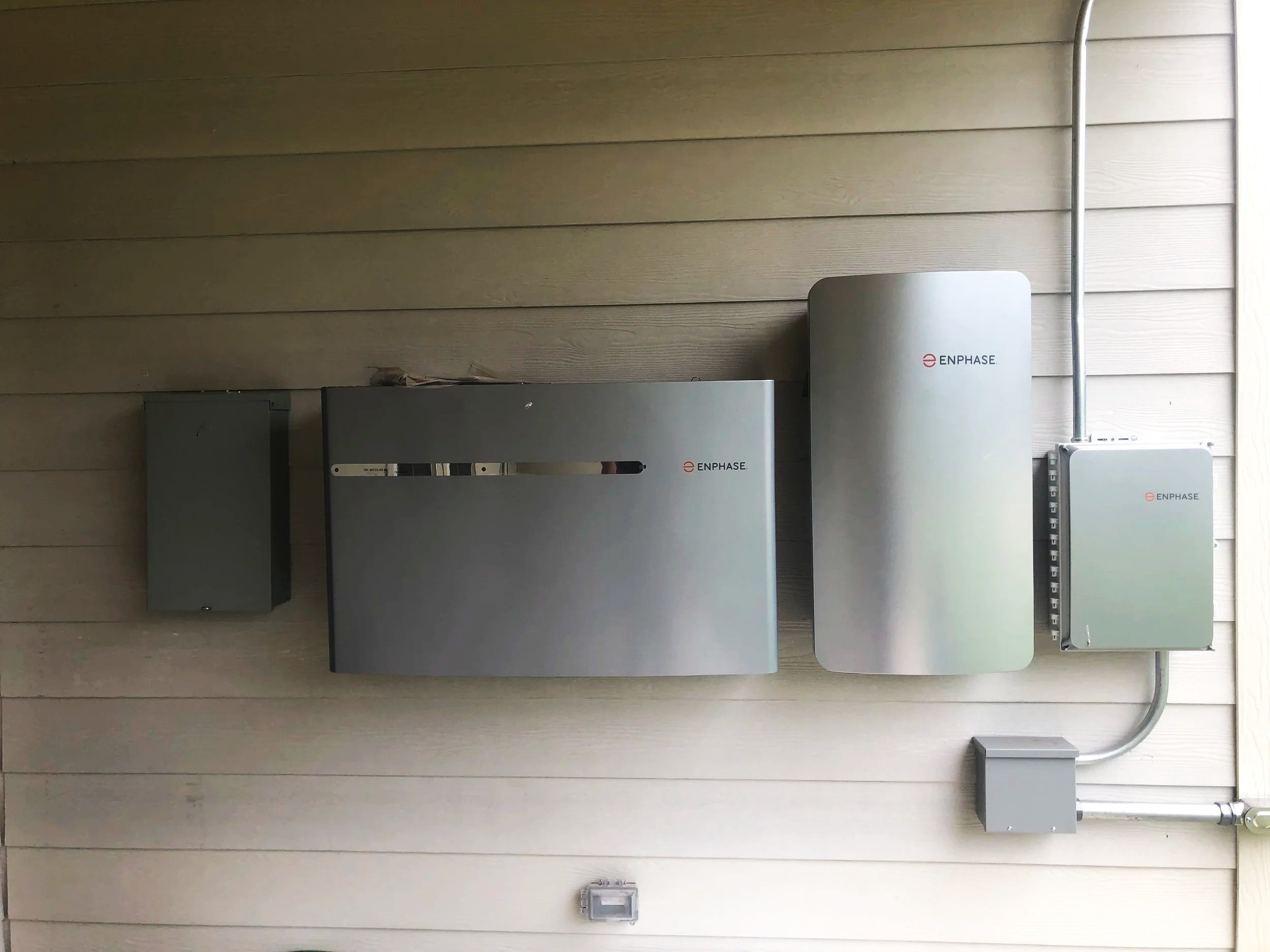 Array of solar energy equipment with Enphase branding mounted on a wooden wall, including invertors and electrical boxes.