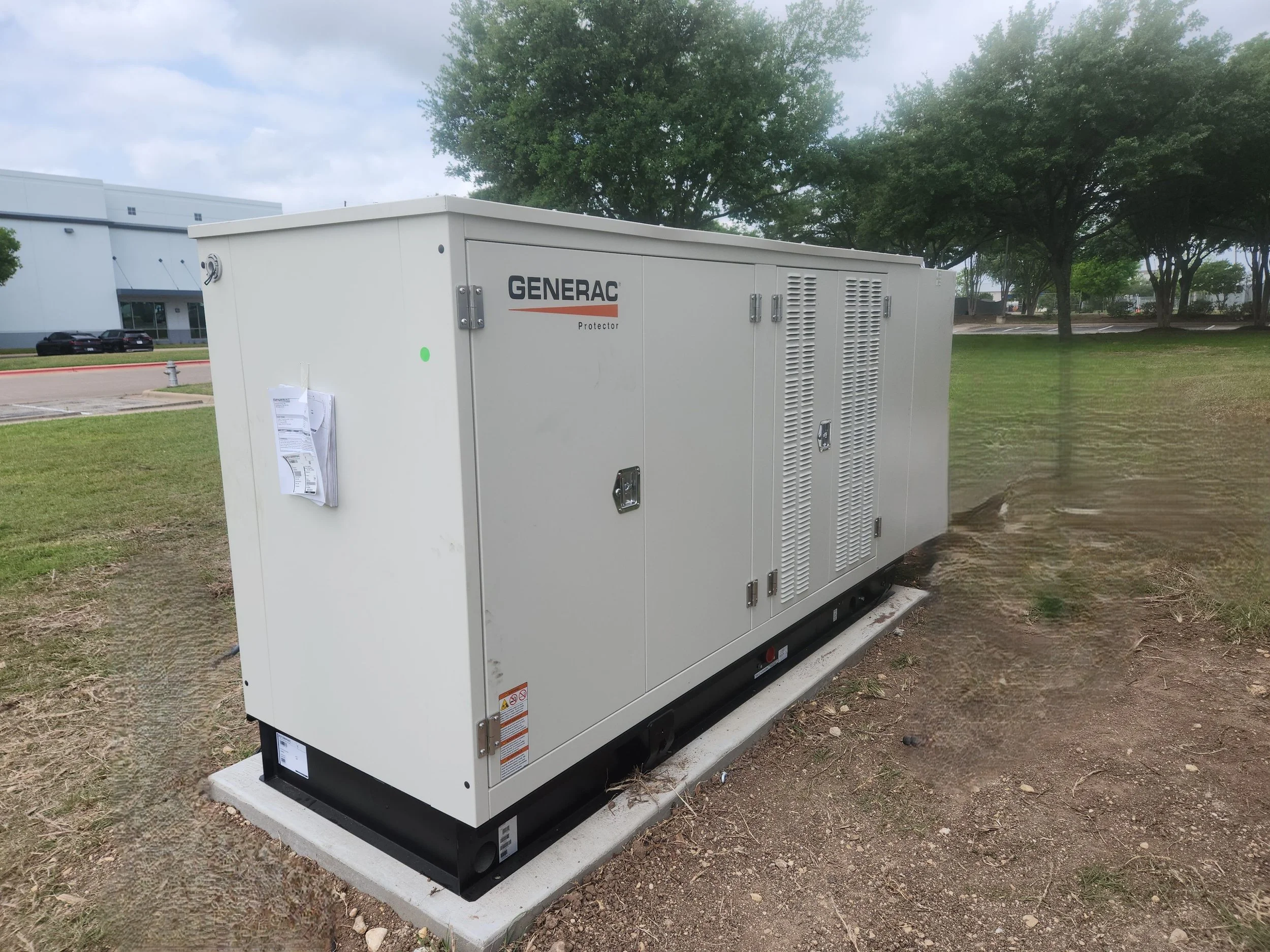 An industrial generator by Generac installed on a concrete pad outdoors near grass and trees.