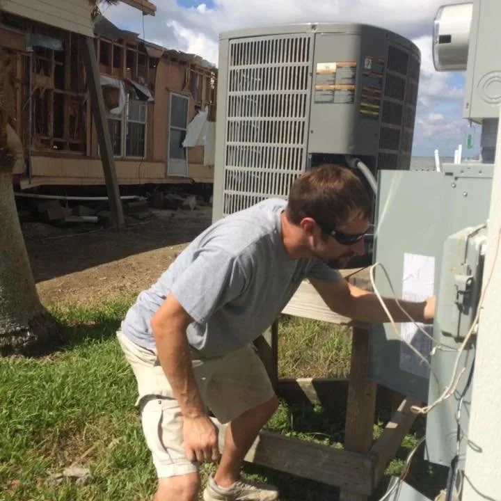 A man wearing sunglasses, a gray t-shirt, and shorts working on outdoor air conditioning or electrical equipment outside on grass.