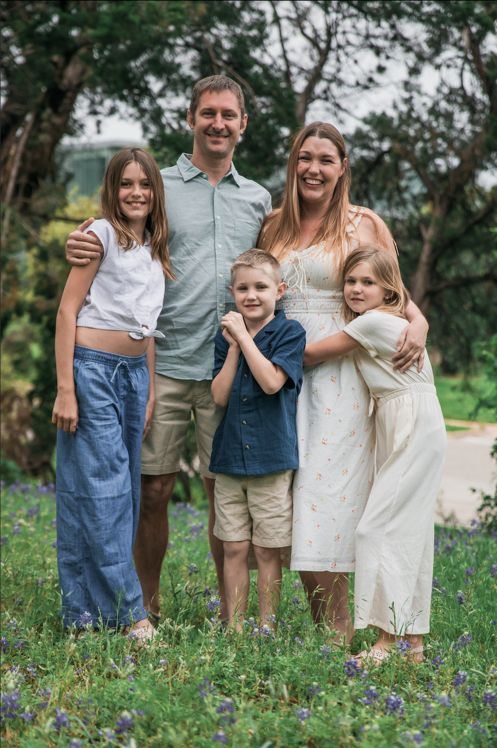 A happy family of five standing outdoors in a park, smiling at the camera. The family includes a father, a mother, and three children, surrounded by green trees and purple flowers.