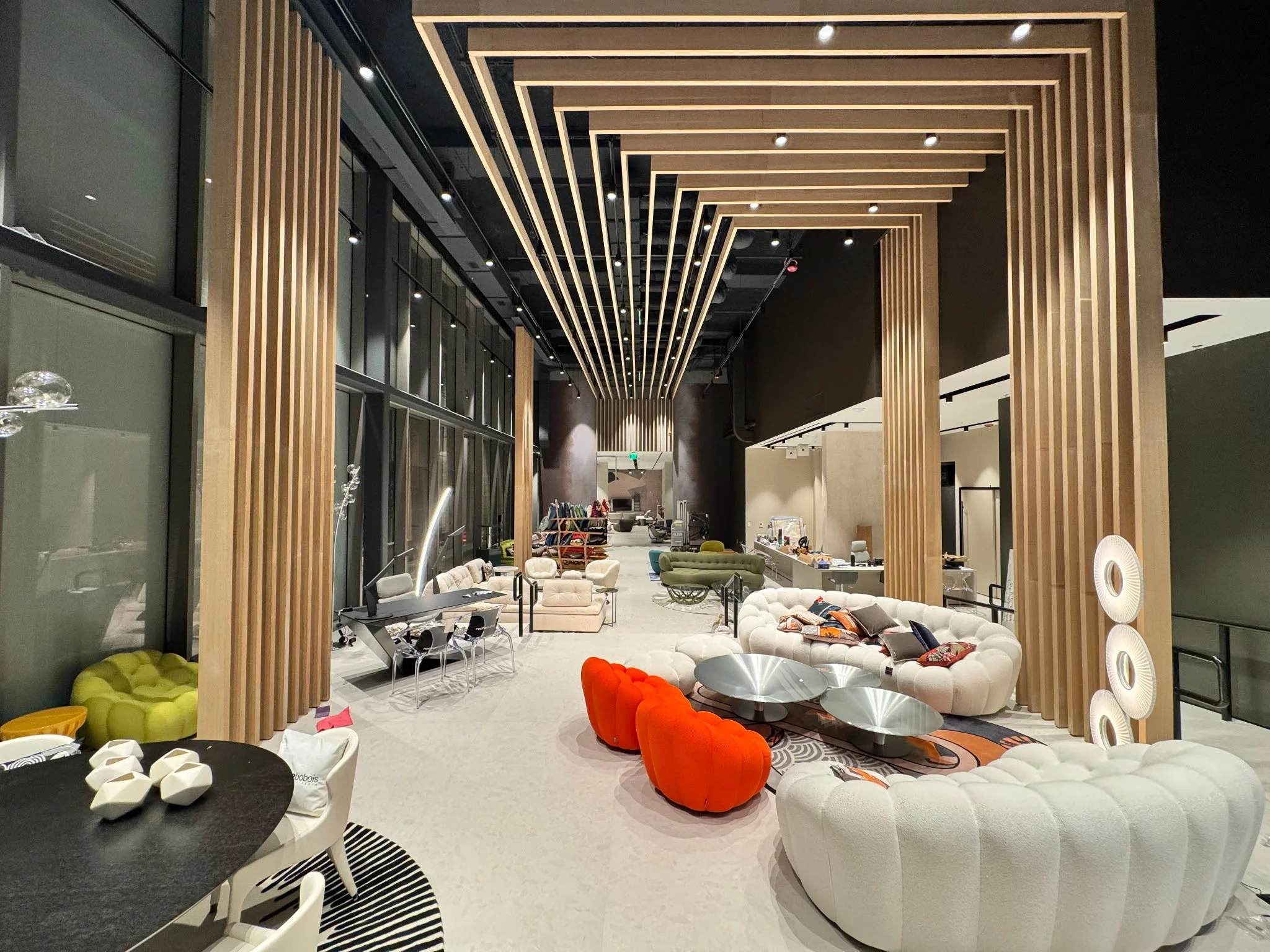 Modern hotel lobby with contemporary furniture, including white and orange sofas, glass coffee tables, and geometric decor. Features wooden slat ceiling design and large windows.