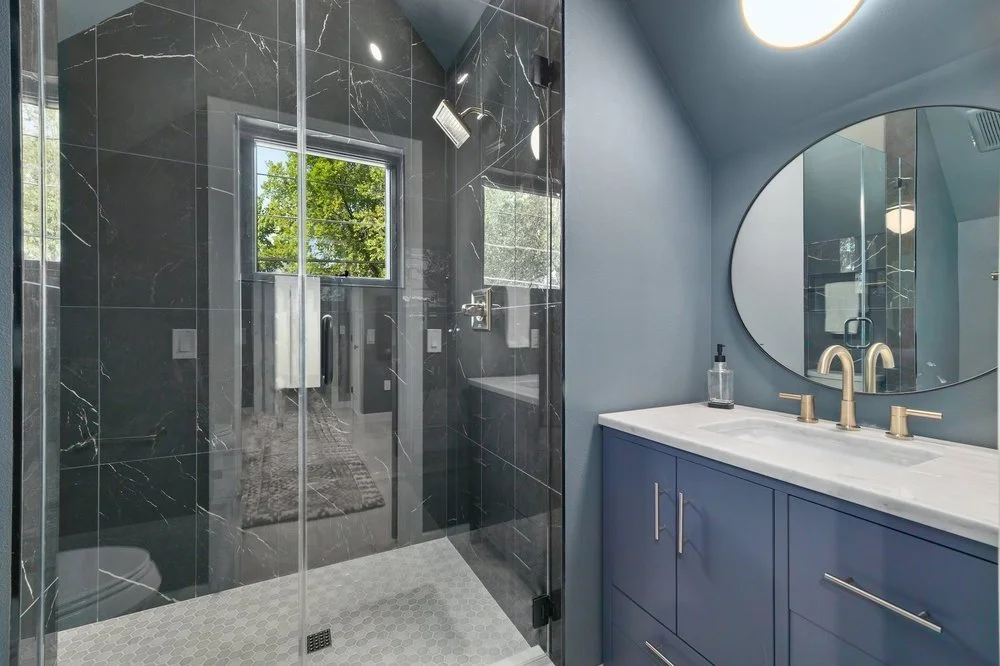 Modern bathroom with a blue vanity, single sink, round mirror, and a glass-enclosed shower with black marble tiles and a window showing green trees outside.