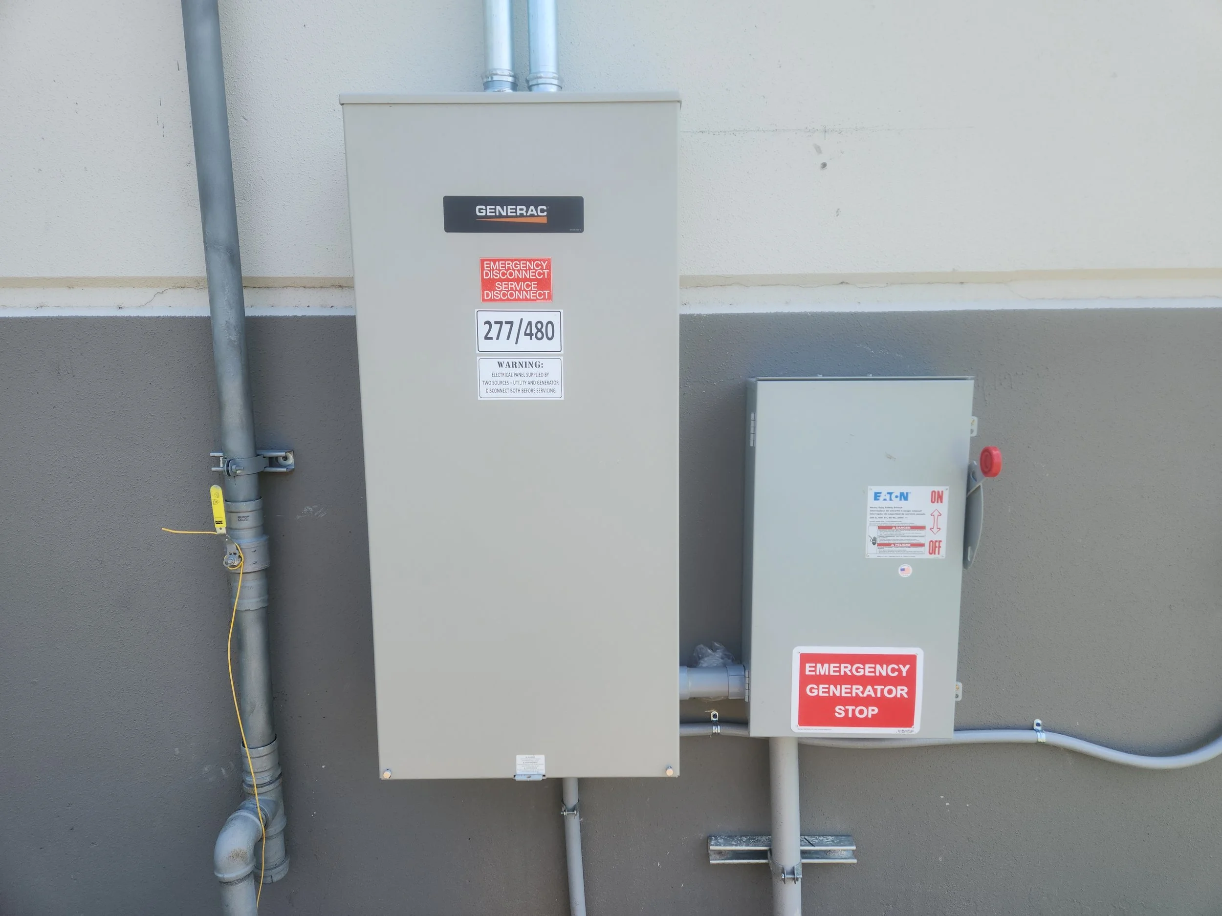 An outdoor electrical power generator and control box mounted on a wall, with warning labels and conduit pipes, for emergency power backup.