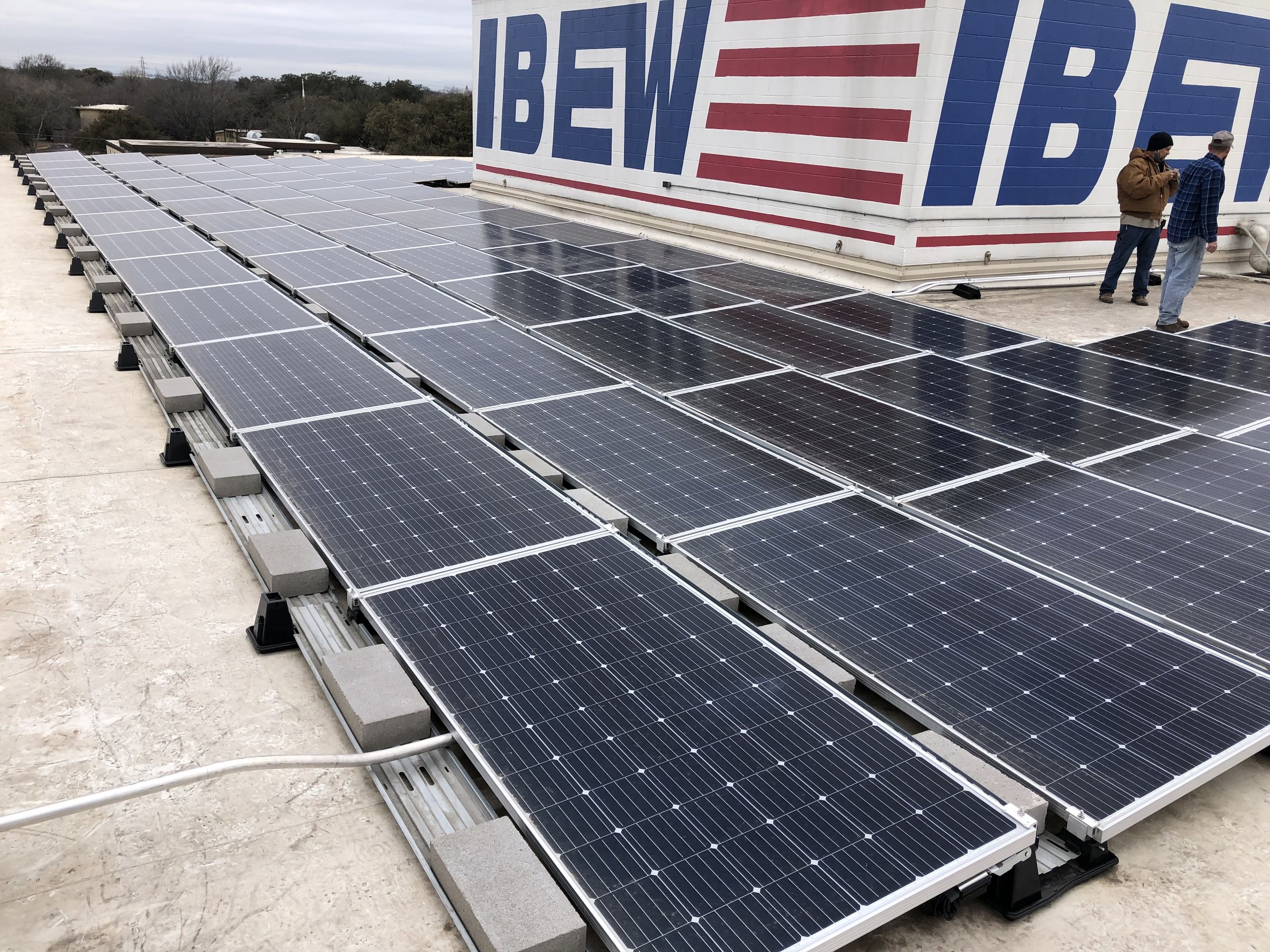 Array of solar panels on a rooftop near a building with a large sign displaying the logo 'IBEW'.