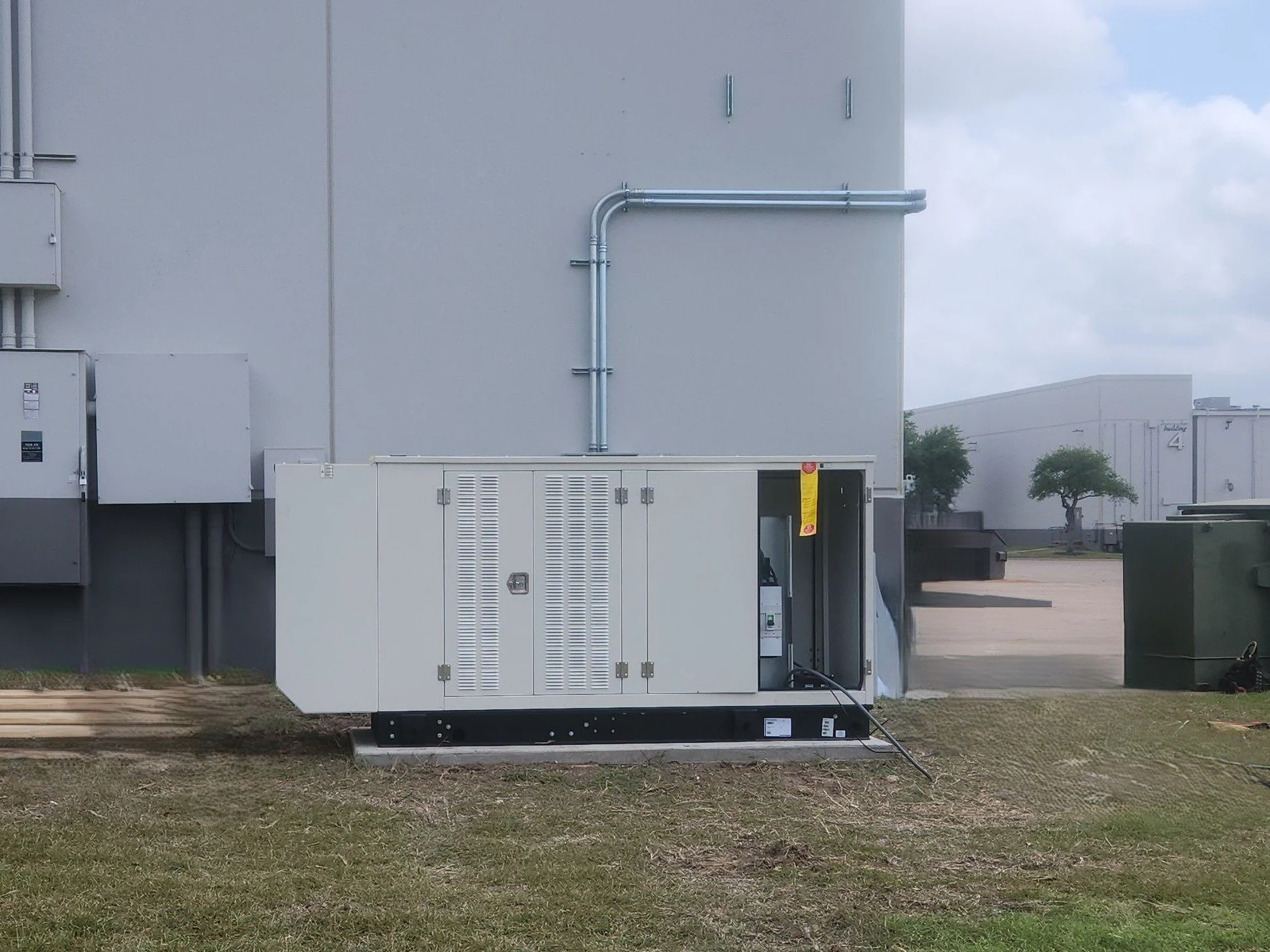 Industrial electrical equipment outside a building with gray walls, pipes, and utility boxes, on a patch of grass and concrete with trees and cloudy sky in the background.