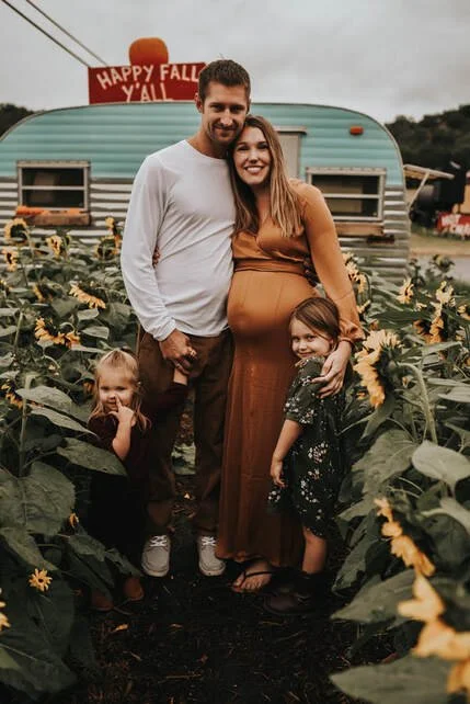 A family of four standing in a sunflower field during fall, with a vintage camper and a sign that says "Happy Fall Y'all" in the background.