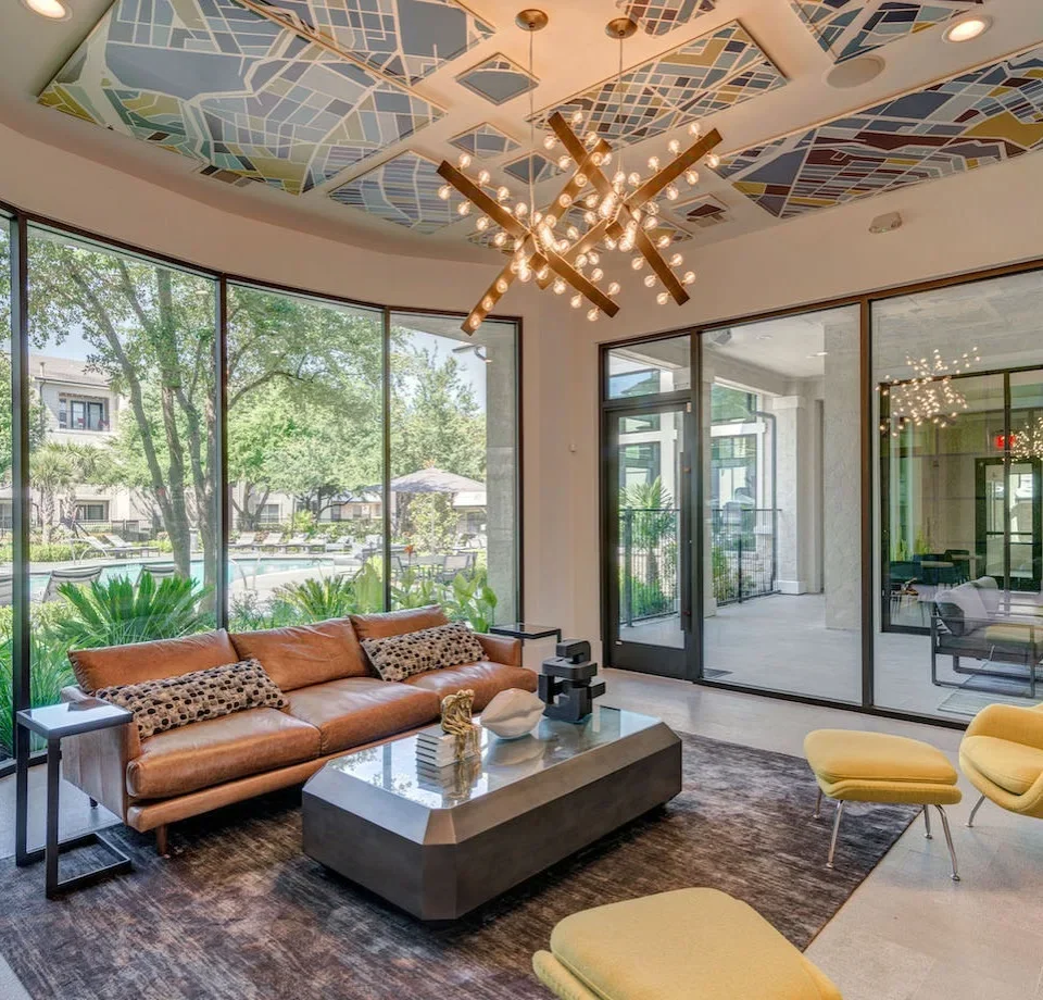 Modern living room with a brown leather sofa, yellow chairs, glass coffee table, large windows showing greenery outside, and a geometric-patterned ceiling with a chandelier.