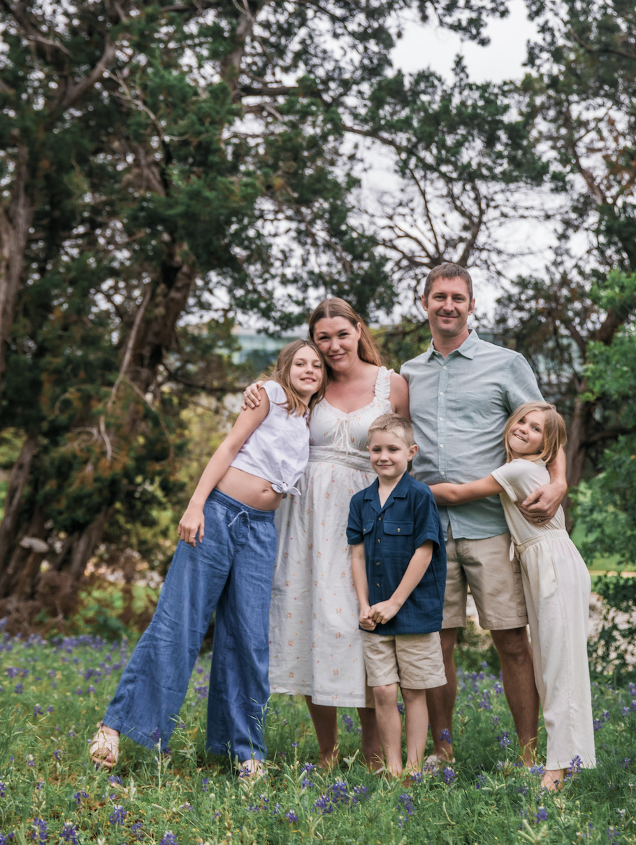 A family of five standing together outdoors in a park-like setting with trees and green grass, smiling and posing for the photo.