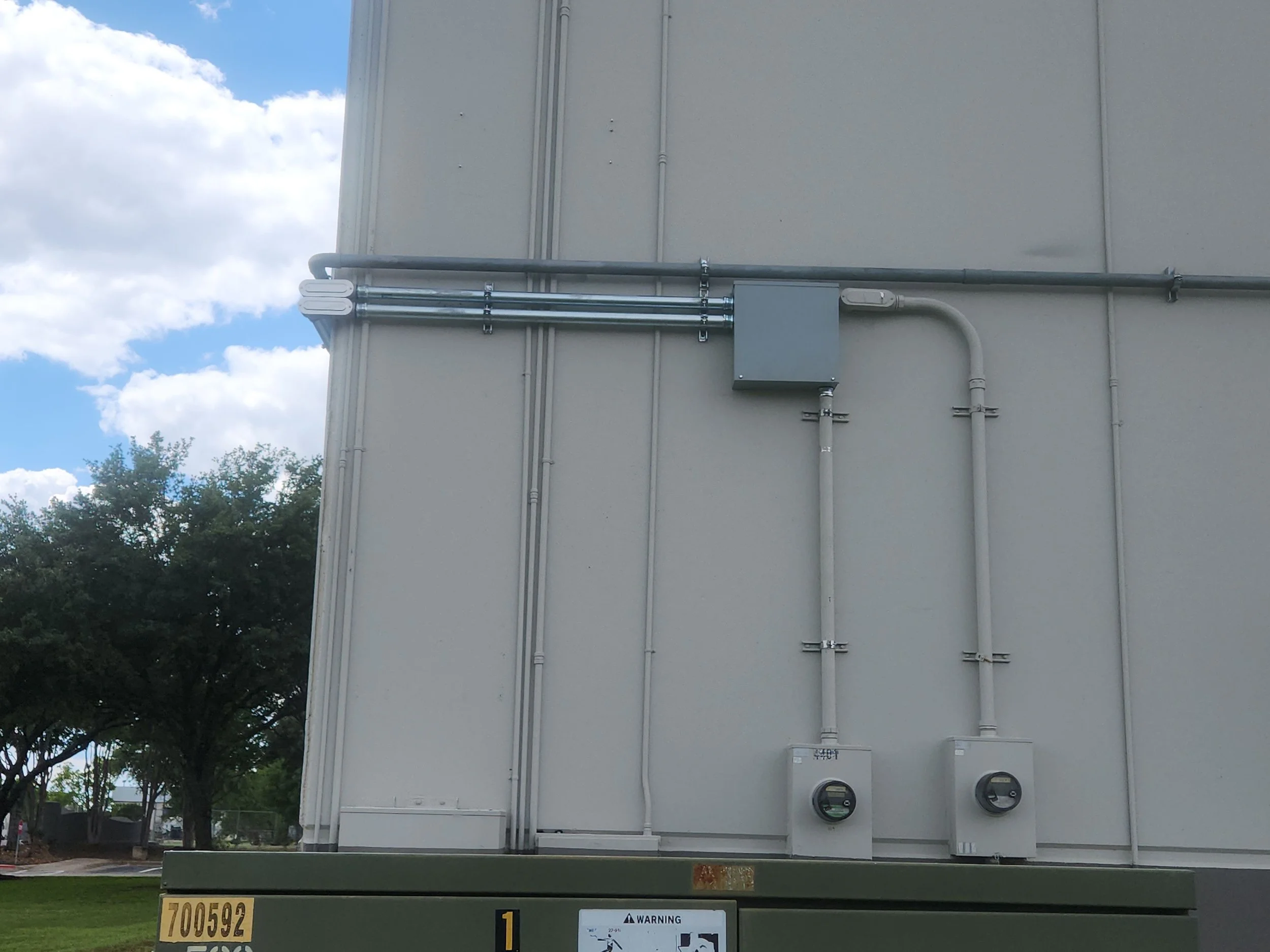 Exterior wall of a building with electrical conduits, a grey electrical box, and two meters mounted on a green base at the bottom of the wall. Trees and a cloudy sky are in the background.