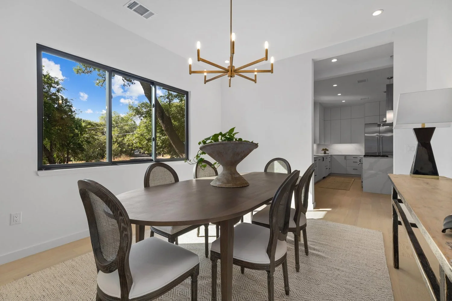 Dining room with wooden table, six chairs, large window showing trees, modern chandelier, and view into kitchen.