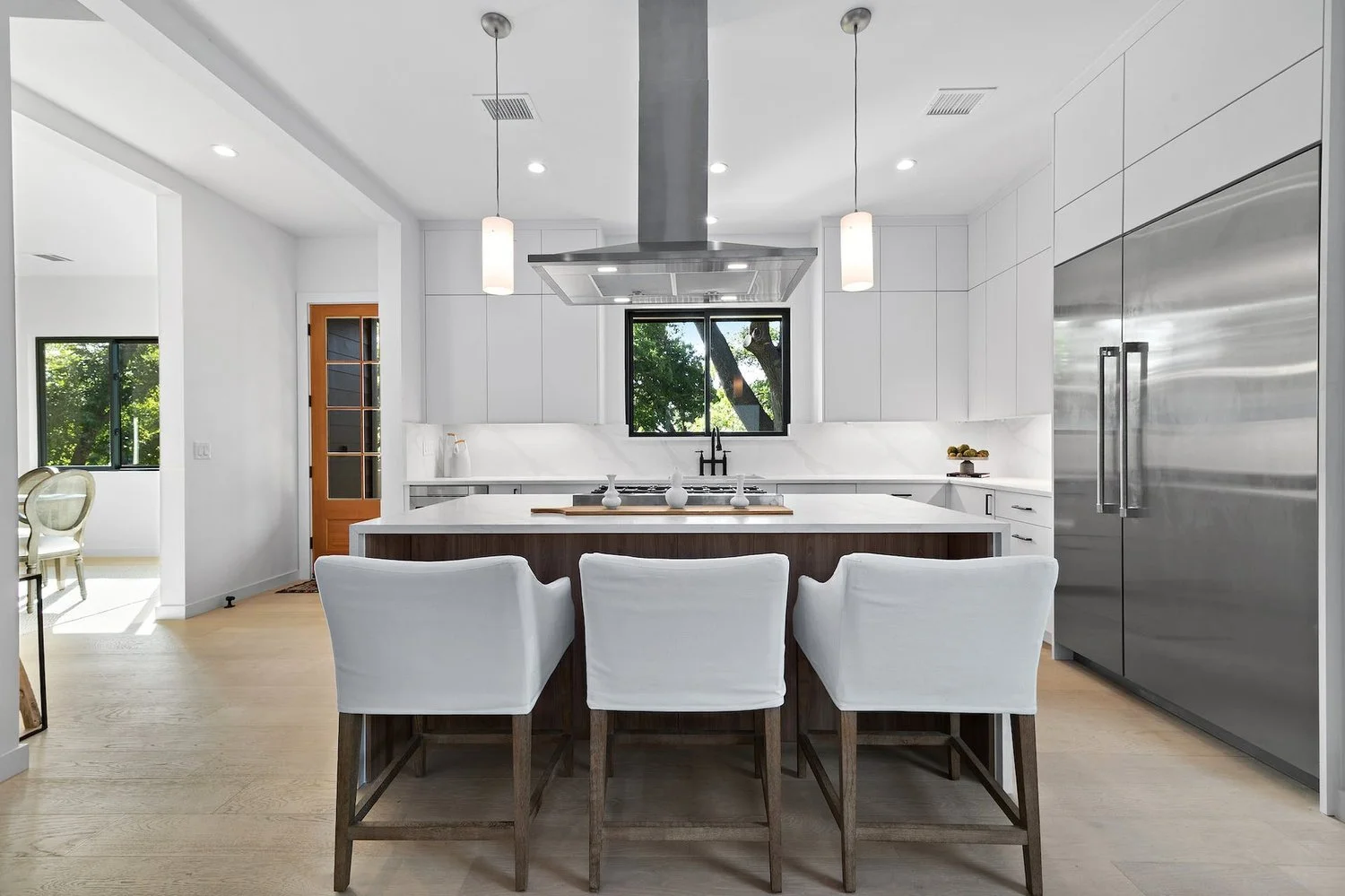 Modern white kitchen with three white chairs at a kitchen island, stainless steel refrigerator, black window showing trees outside, and white cabinetry.