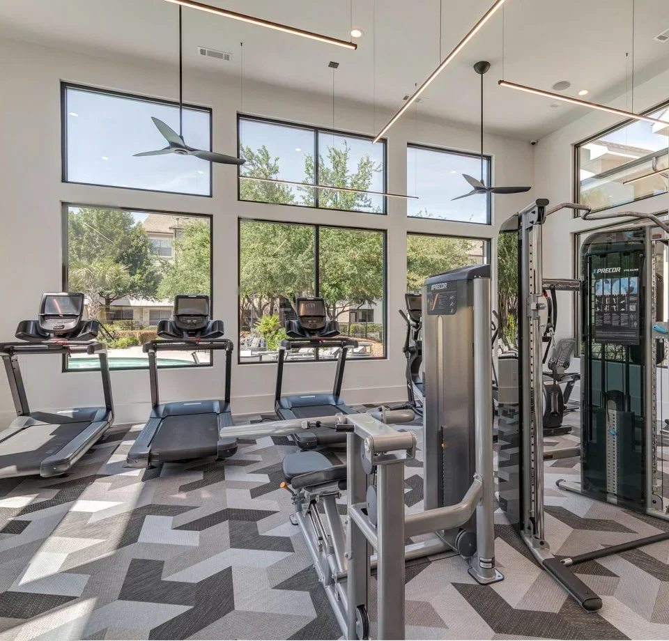Modern gym with treadmills, cardio machines, and weight equipment, large windows with a view of trees outside, ceiling fans and contemporary lighting.