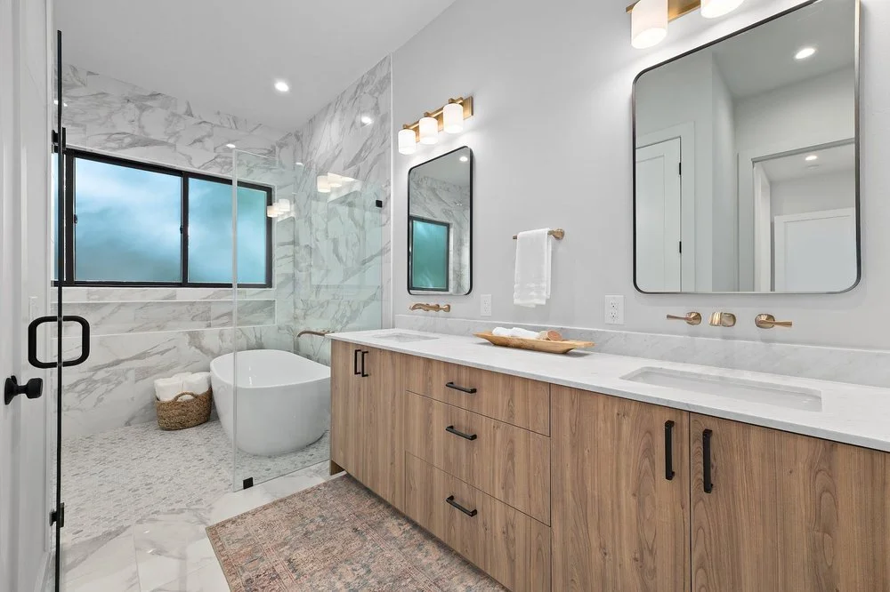 Modern bathroom with a wooden vanity, a large mirror, a bathtub near a window, marble walls, a glass shower enclosure, and a patterned rug on the floor.