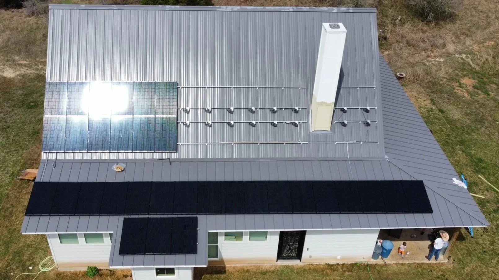 A house with a metal roof featuring solar panels and a solar water heater, with people and outdoor items on the porch.