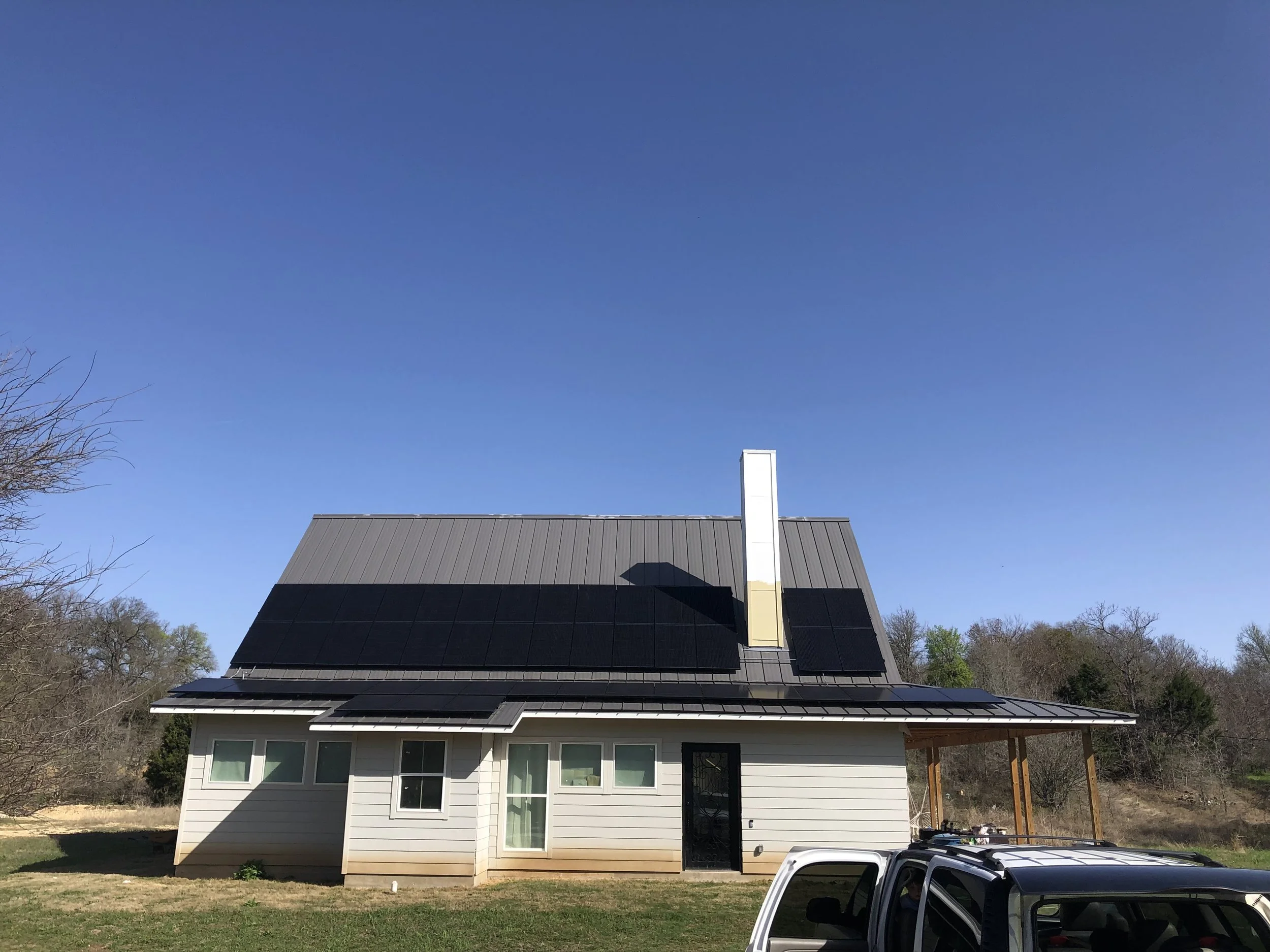 House with solar panels on roof, blue sky, trees in background, vehicle in foreground.