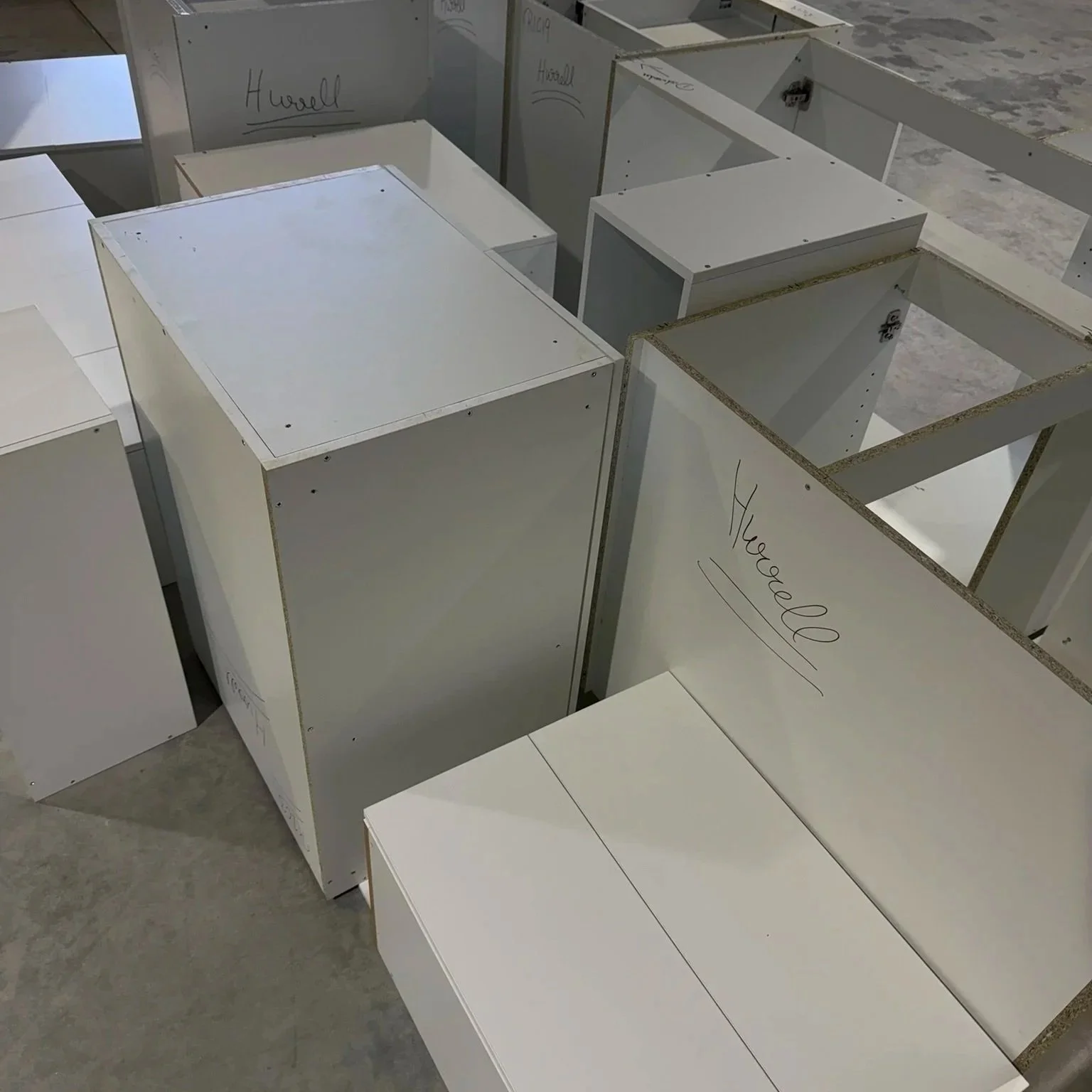 Multiple white furniture parts, some labeled with names like "Howell" and "Yhuegel" written in black marker, arranged on a concrete floor, possibly for assembly or sale.