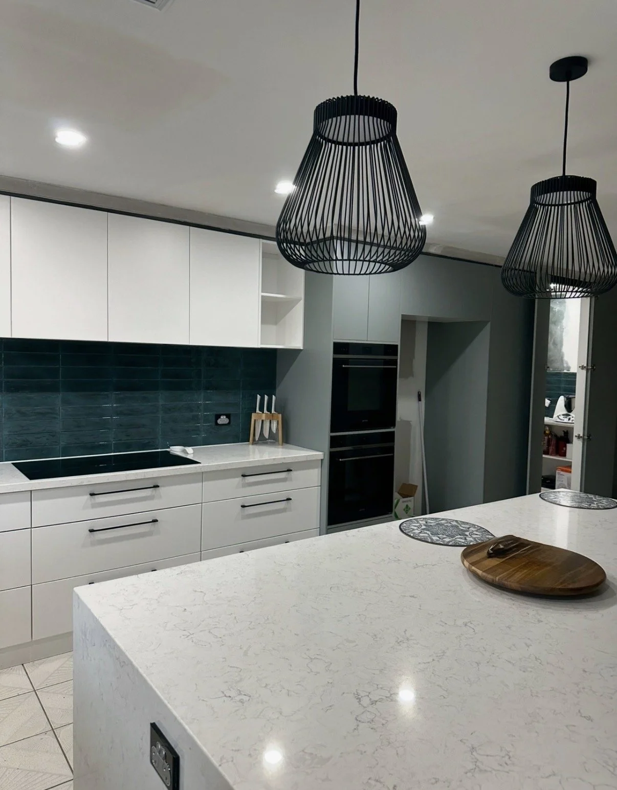 Modern kitchen with white cabinets, teal tile backsplash, and black pendant lights over a white marble island.