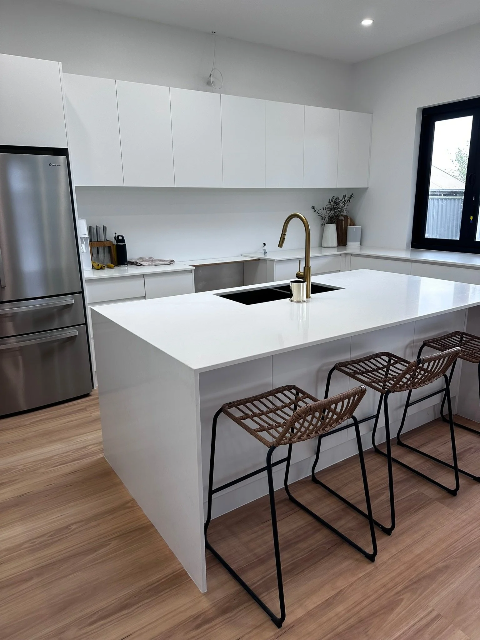 Modern kitchen with a white island counter, three wicker and metal bar stools, a gold faucet, white cabinetry above, and a large window.