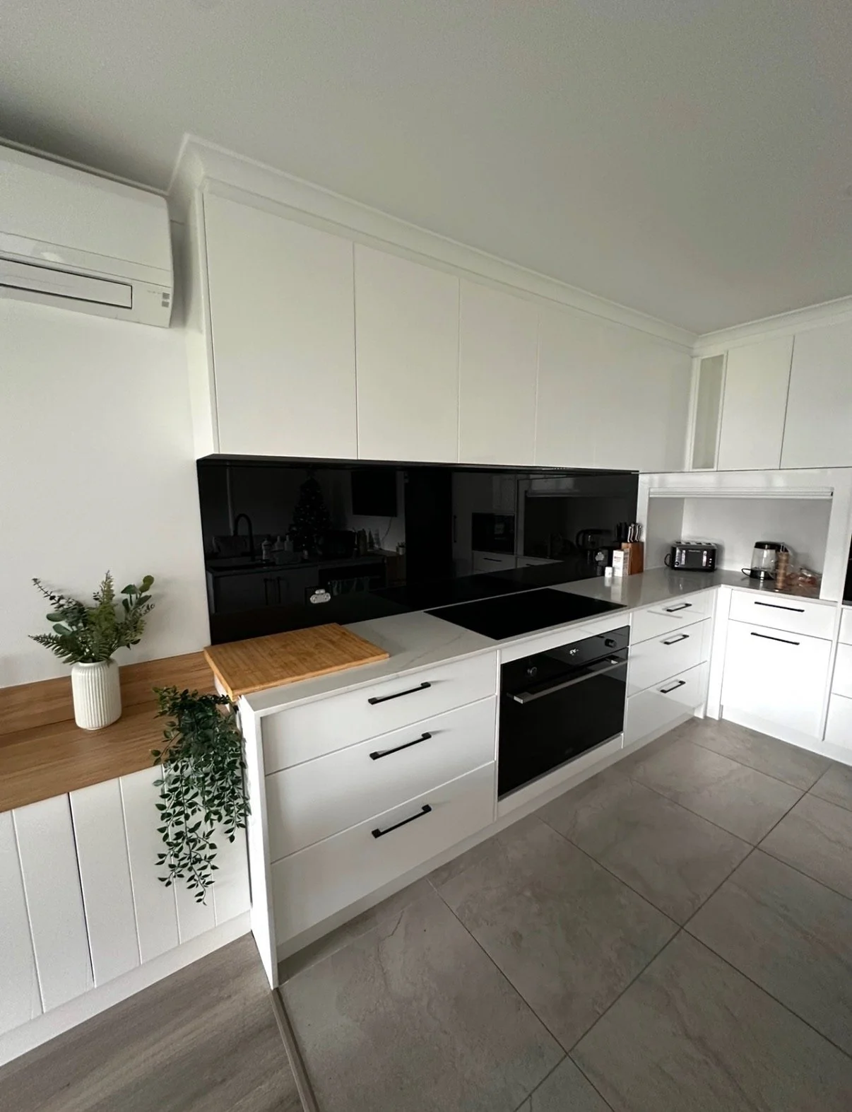 Modern white kitchen with black appliances and gray tiled flooring, decorated with green plants and wooden accents.