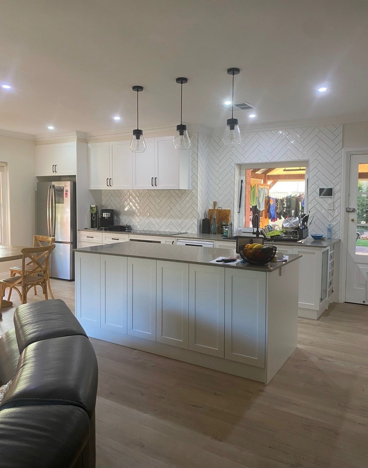 Modern white kitchen with island, hanging pendant lights, stainless steel refrigerator, and view of outdoor laundry area.