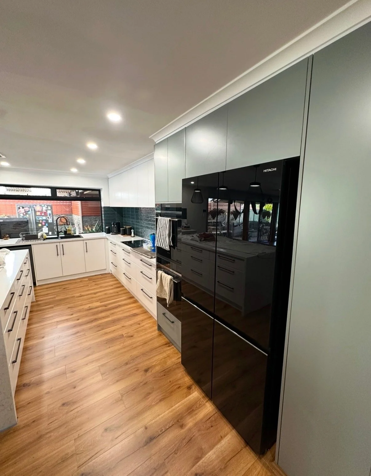 Modern kitchen with white cabinets, black appliances, wooden flooring, and large windows with outdoor view.