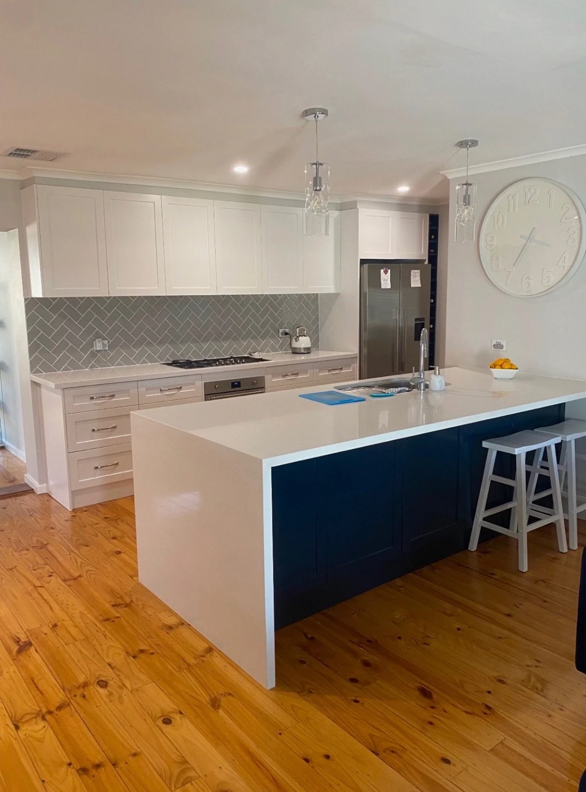 Modern kitchen with white walls and upper cabinets, gray backsplash in a herringbone pattern, wooden floors, and a large island with a white countertop and dark blue base. There are two stools at the island, a stainless steel refrigerator, a stove, and minimal decor.