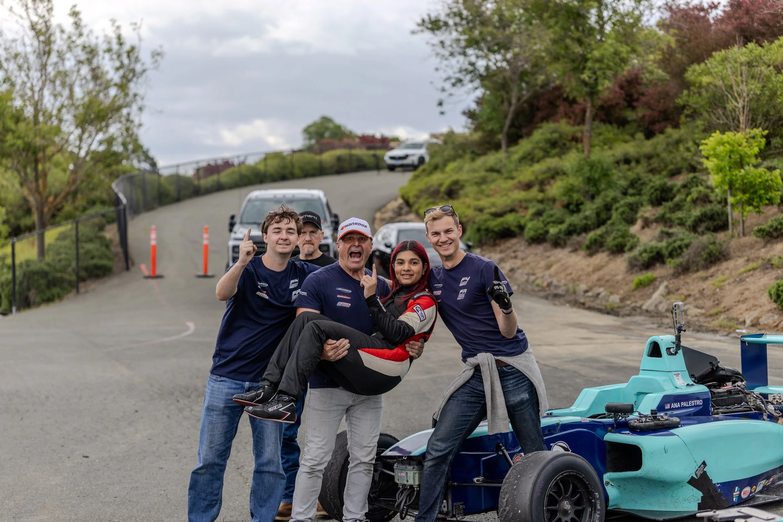 Group of five people, including a man in racing gear, posing with a race car at a race track surrounded by greenery.