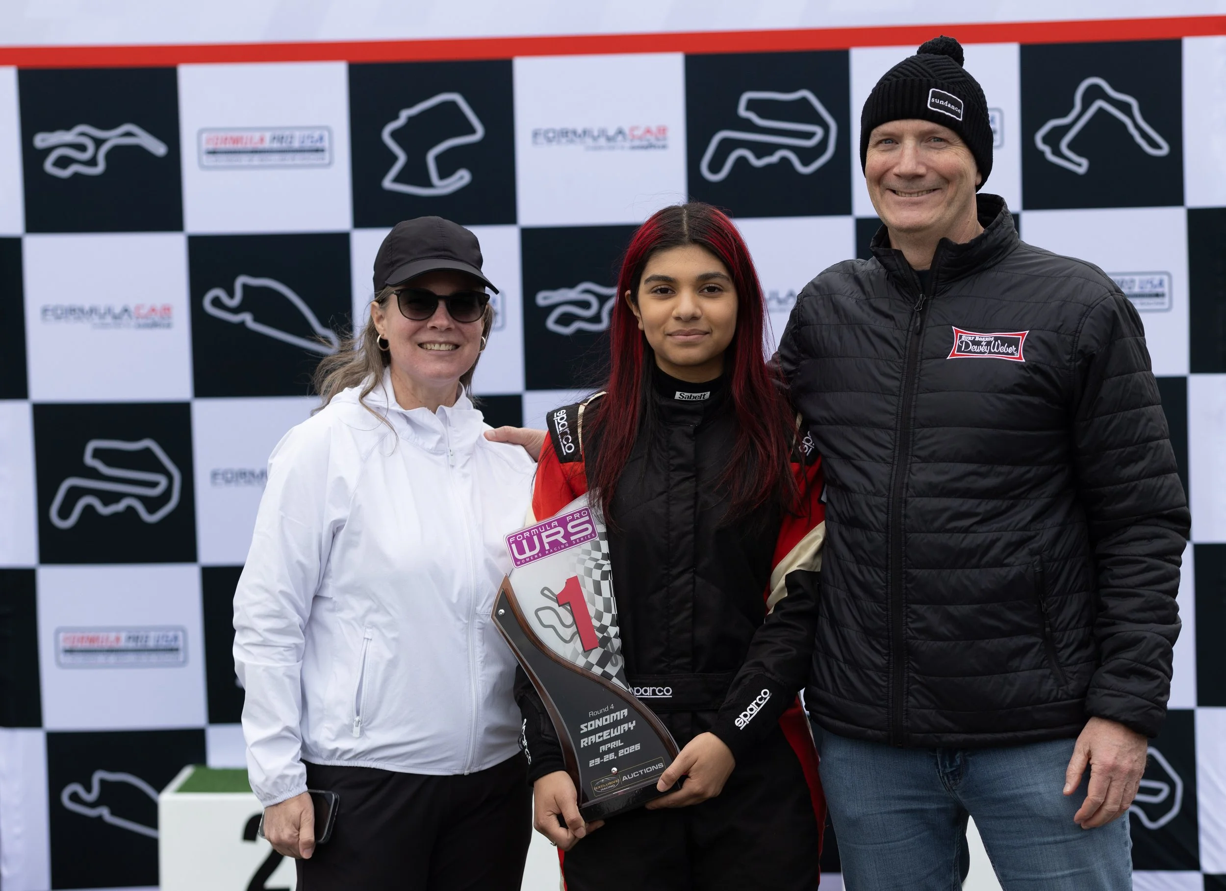 Three people standing on a racing podium in front of a black-and-white checkered backdrop with racing logos. One woman on the left is wearing sunglasses and a white jacket, a young woman in the middle has red hair and is dressed in racing gear, and a man on the right is dressed in a black jacket and knit cap, smiling.