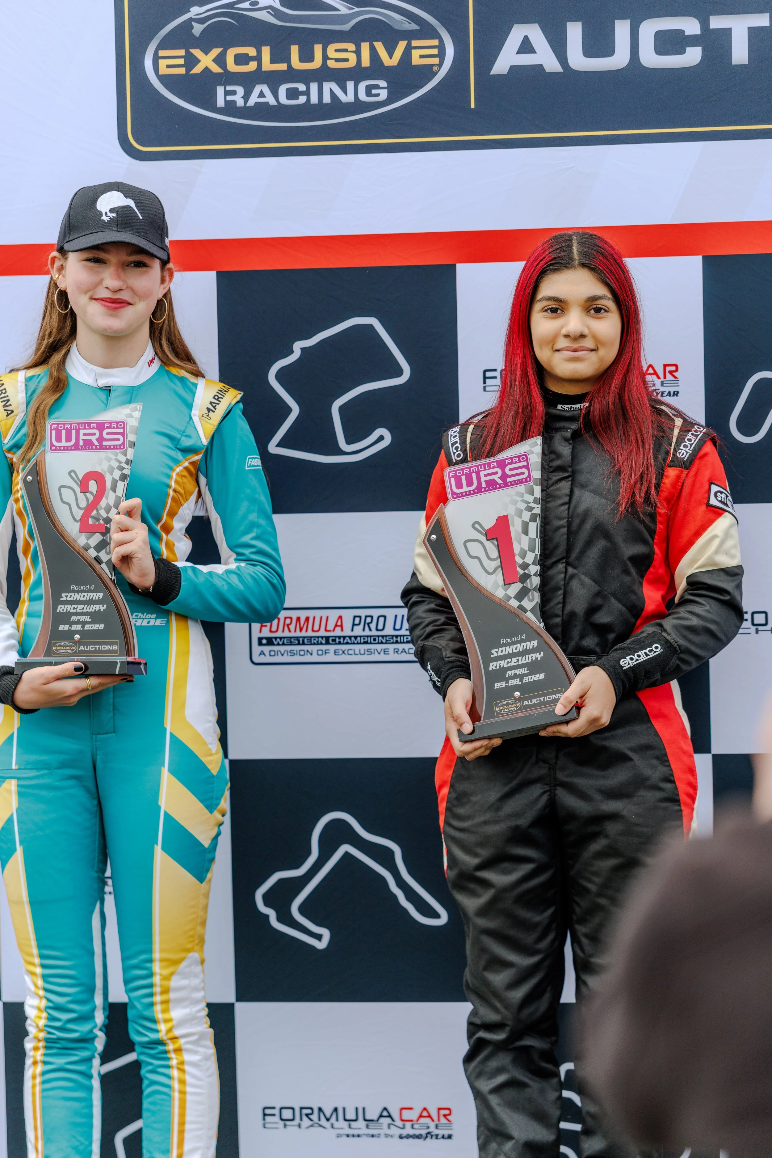 Two young women in racing suits holding trophies at a podium event for Formula Pro WRS at Sonoma Raceway, with a backdrop featuring racing logos and graphics.