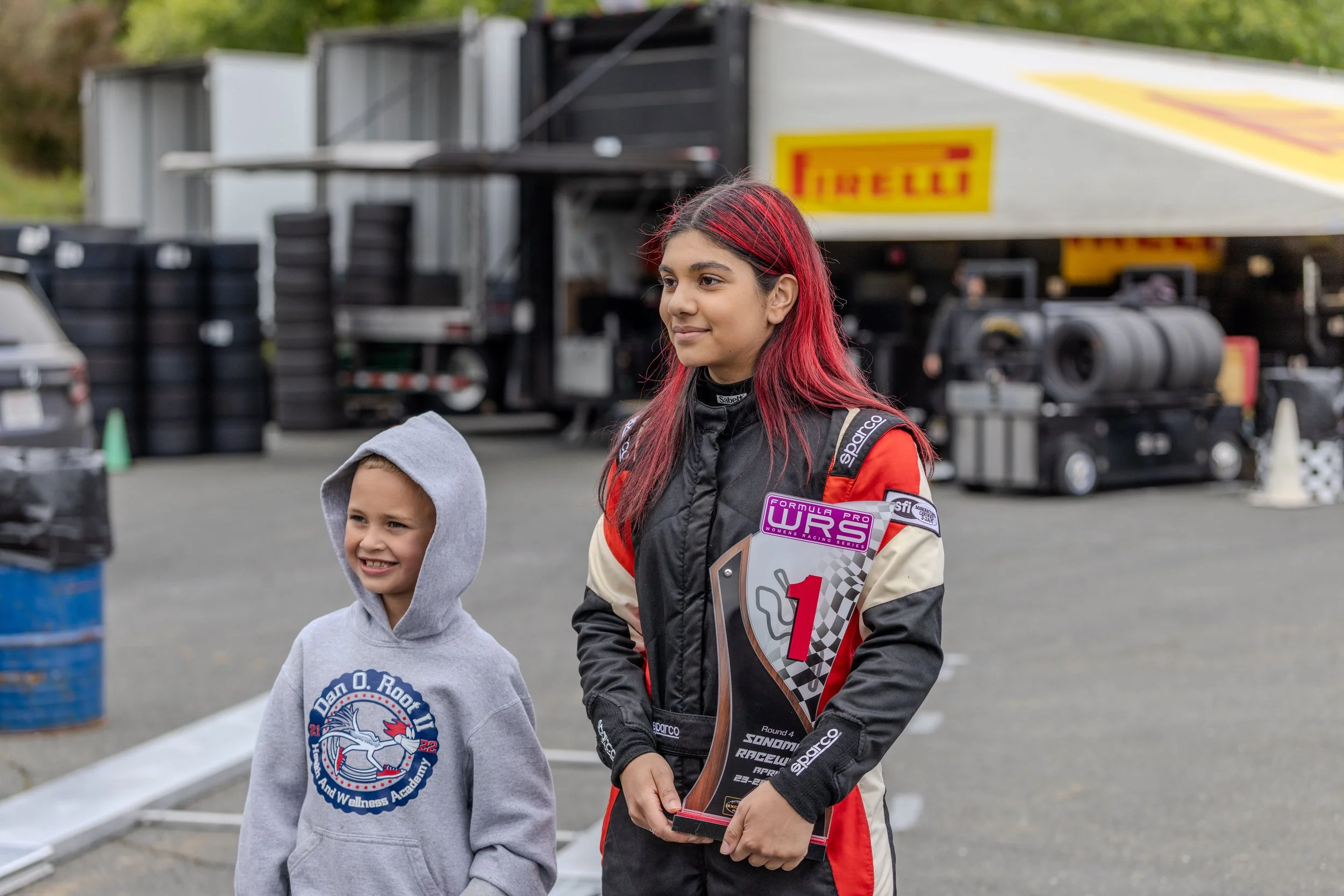 A young woman radiates happiness as she holds a trophy, standing next to a smiling young boy. They are outdoors at a racing event with a black tire stack and a trailer with Pirelli branding in the background.